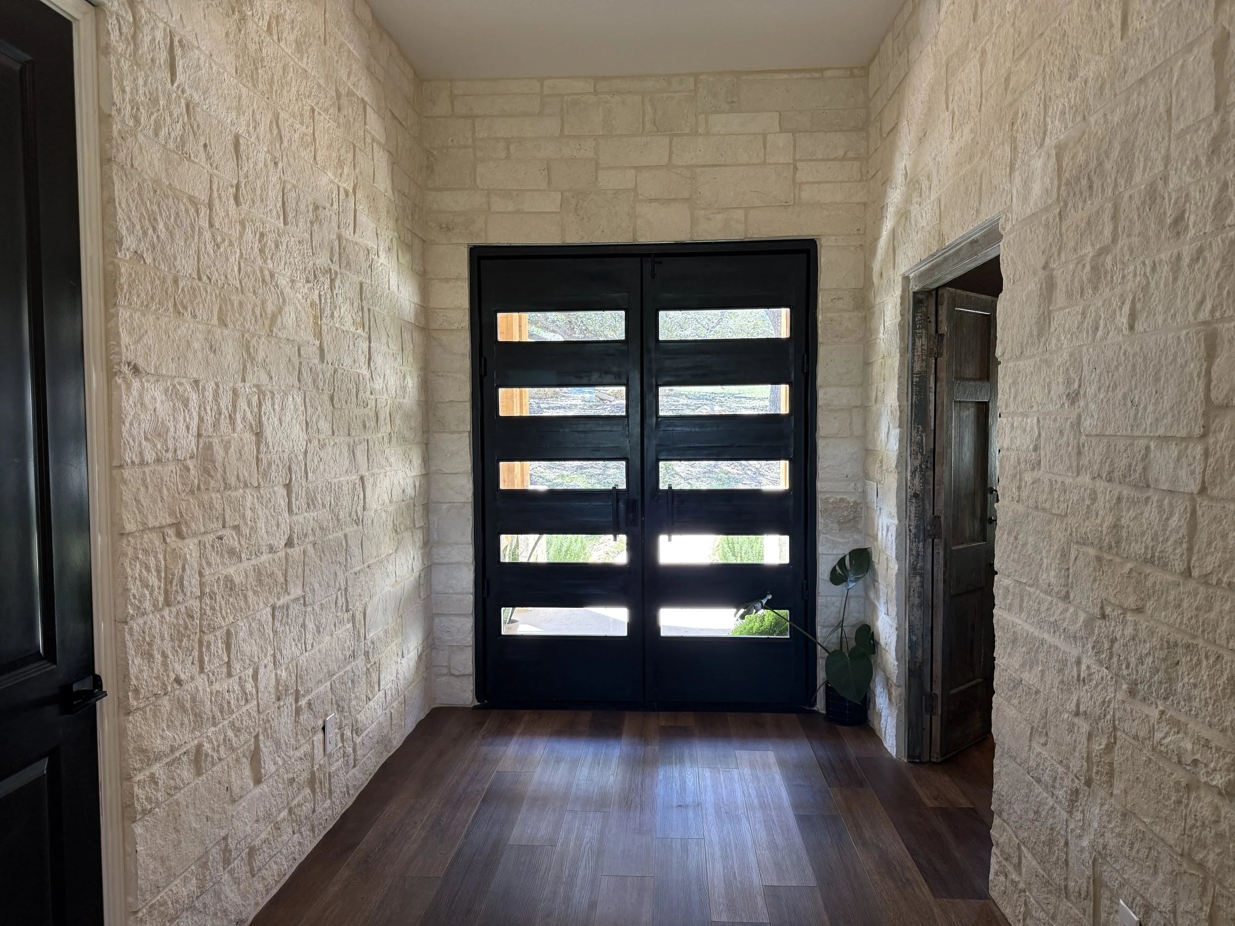 Interior entryway with black modern double doors with horizontal glass panels, stone walls, hardwood floor, and a potted plant near the door.