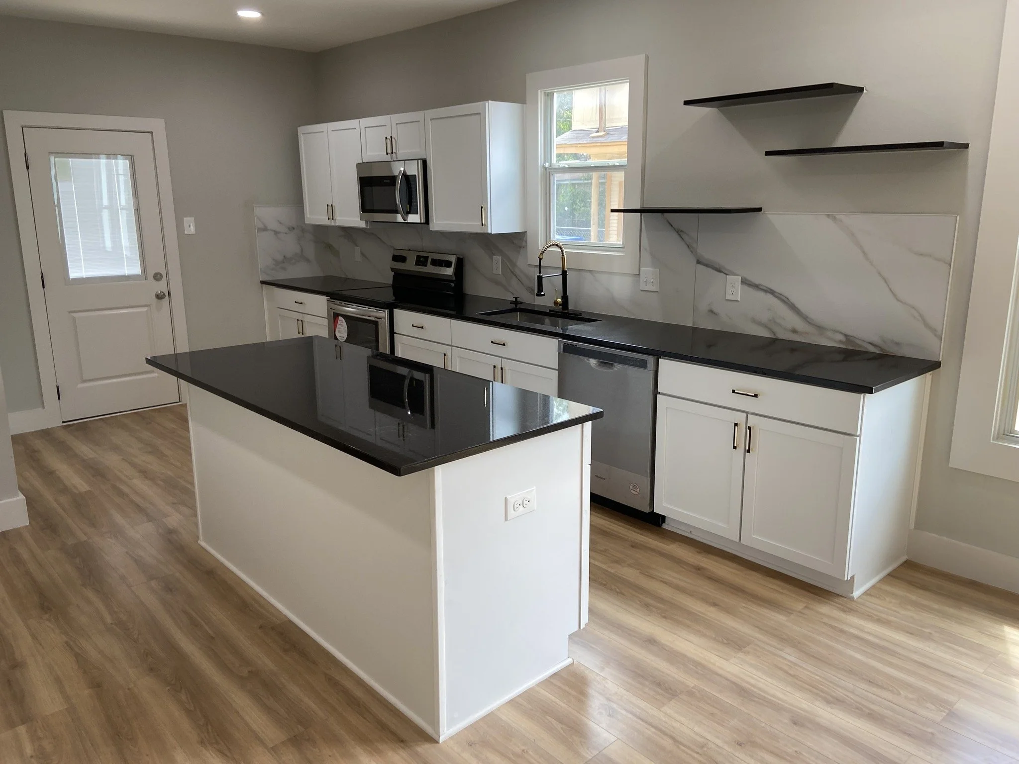 Modern kitchen with white cabinets, black countertops, marble backsplash, stainless steel appliances, wood flooring, and open shelves.