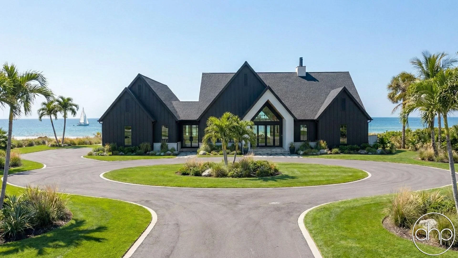 Black modern house with steep roof, large glass front entrance, surrounded by well-manicured lawn and tropical plants, with the ocean and sailboat in the background.