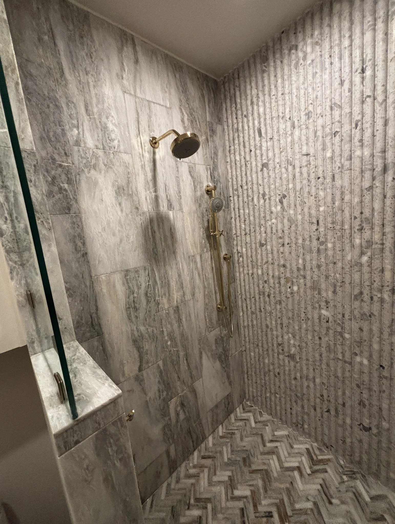 A shower with gold fixtures, including a large overhead shower and a handheld shower, set against a wall with a mix of gray marble and ribbed concrete textures, and a patterned tile floor.
