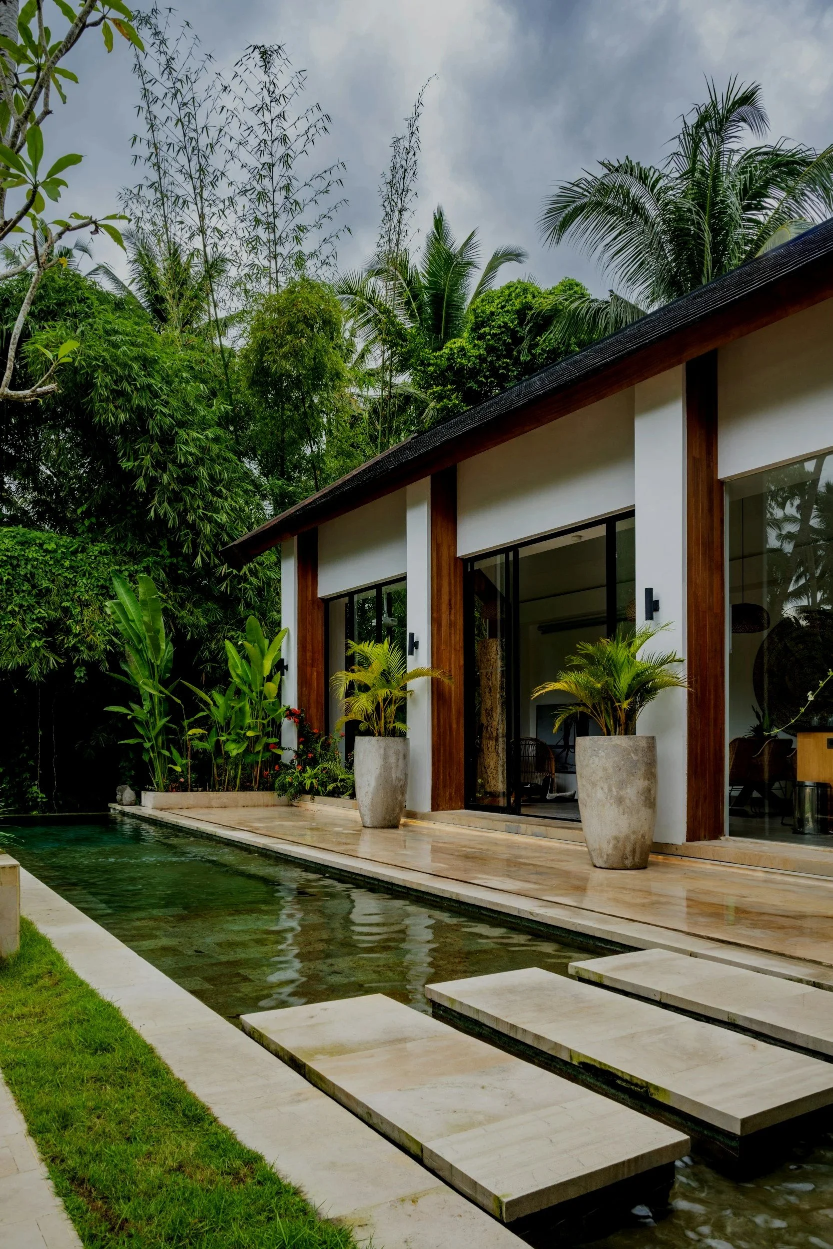 Modern house with sliding glass doors, surrounded by lush tropical plants, with a narrow reflecting pool and stepping stones leading to the house in a tropical garden.