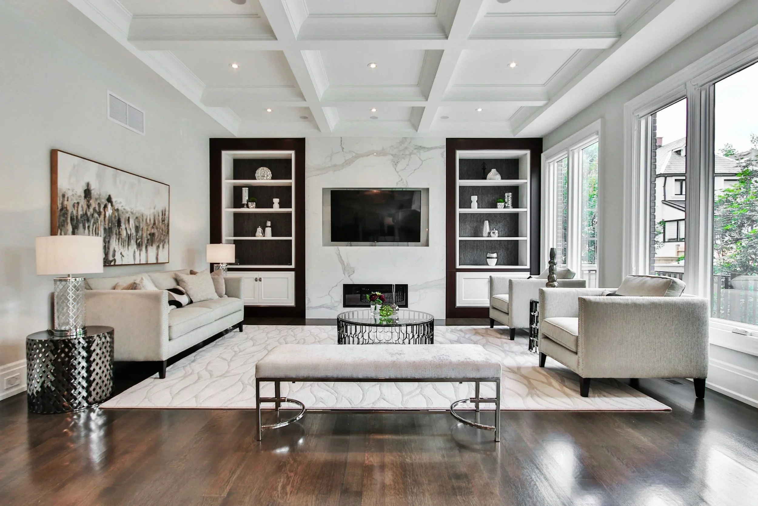 Elegant living room with white walls, dark hardwood floors, and large windows. Features a white sofa with pillows, two white armchairs, a bench, and a round coffee table. Built-in shelves flank a wall-mounted TV above a fireplace with a marble surround.