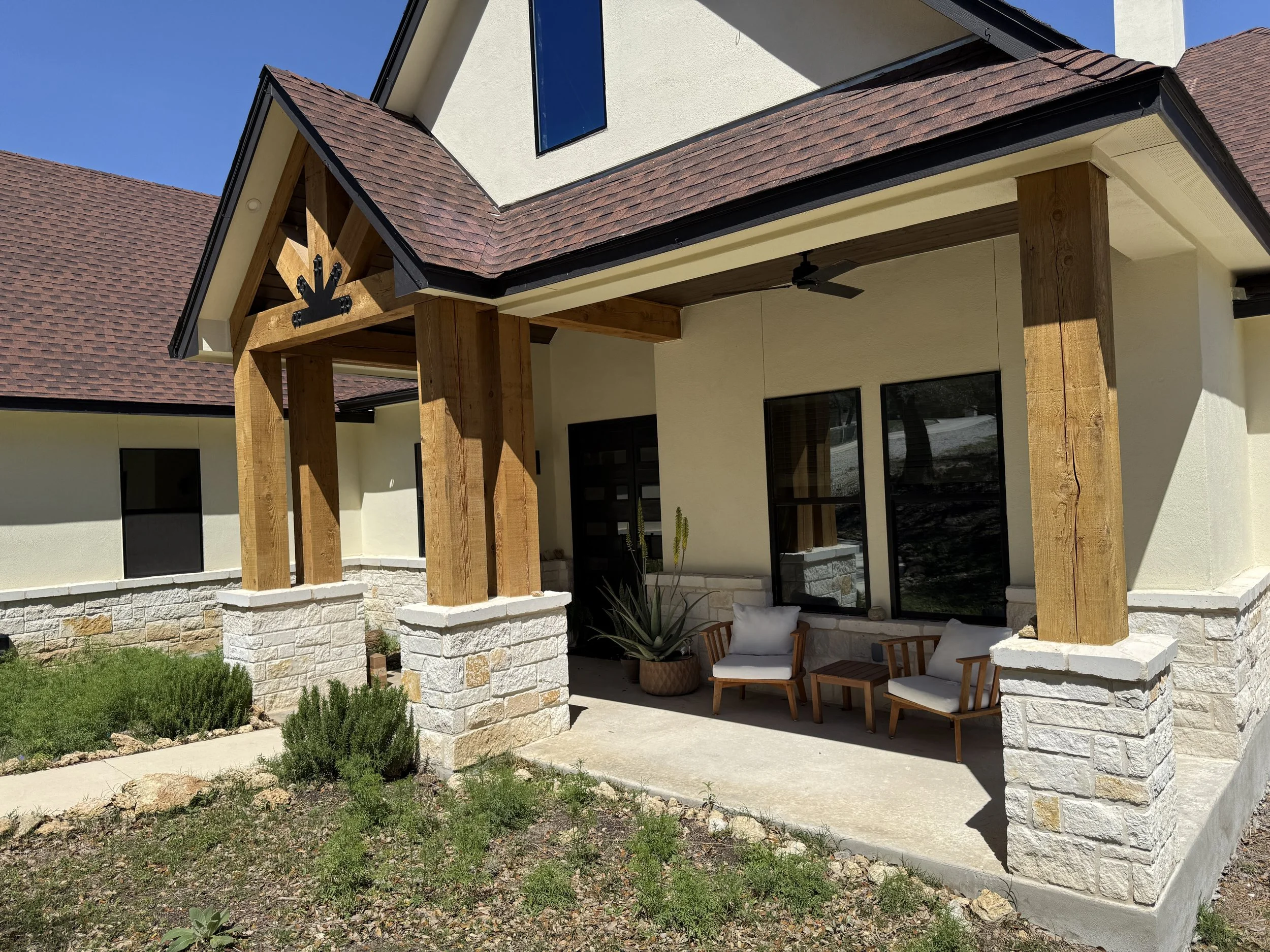 Front porch of a house with a gabled roof, stone pillars, and wooden posts, featuring outdoor chairs and plants, under a clear blue sky.