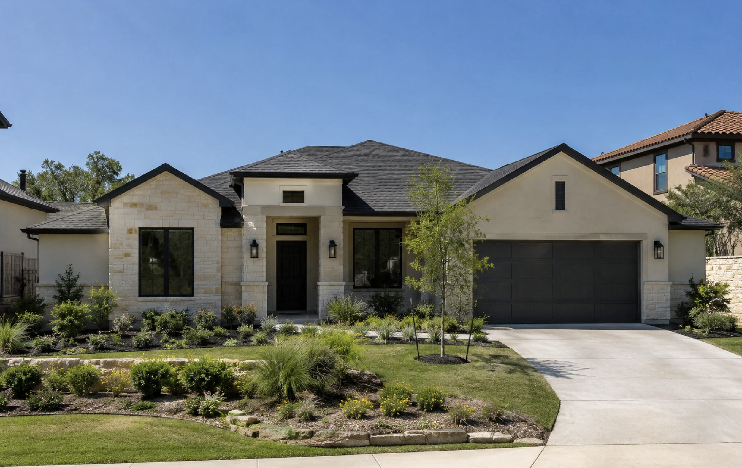 Front view of a modern house with a stone and stucco exterior, dark front door, large windows, a gray garage door, and landscaped front yard with a walkway and small trees, under a clear blue sky.