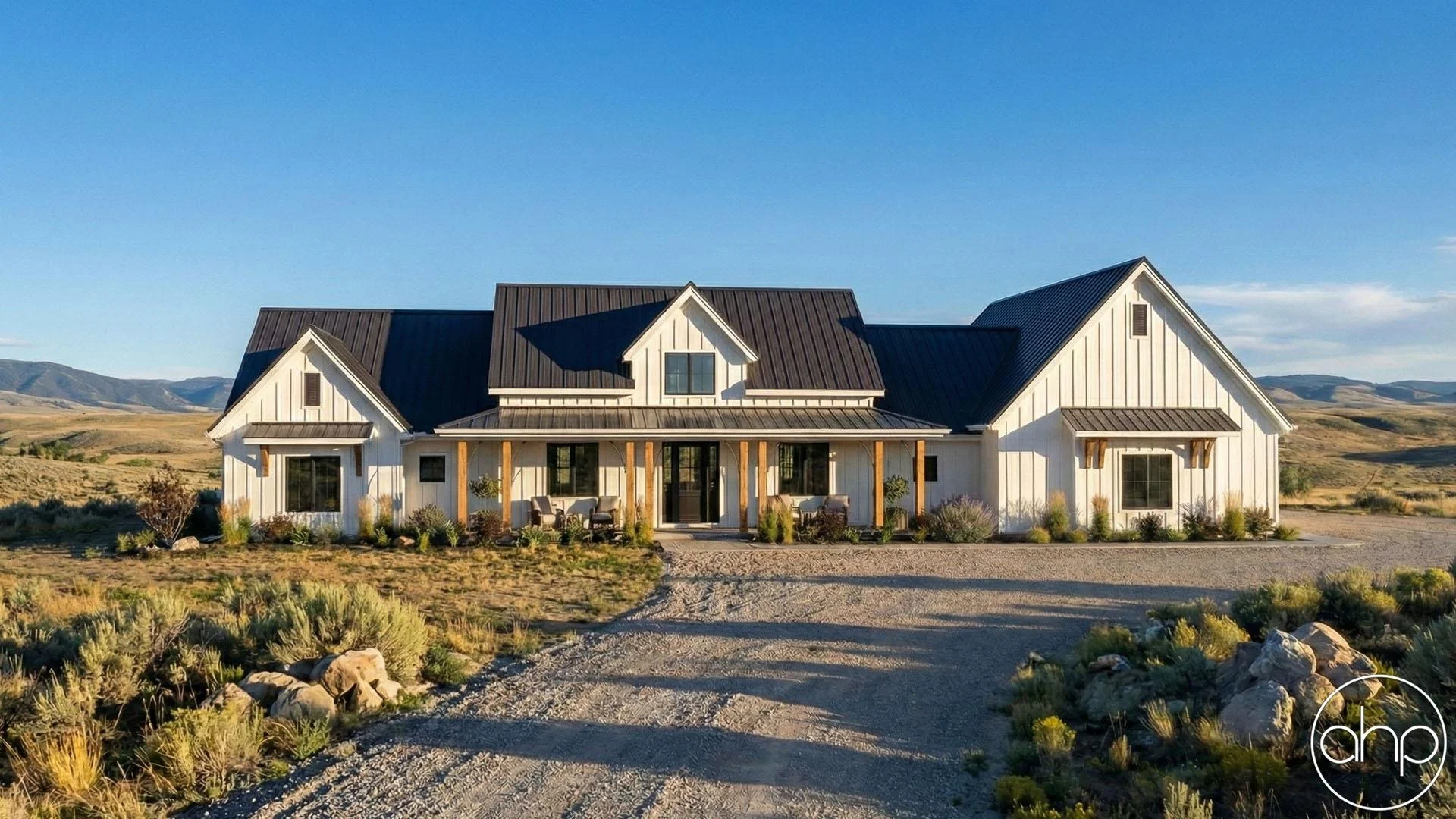 A modern white farmhouse with a black metal roof, situated in a rural landscape with mountains in the background. The house has a front porch with wooden supports and outdoor seating, surrounded by shrubs and gravel driveway.