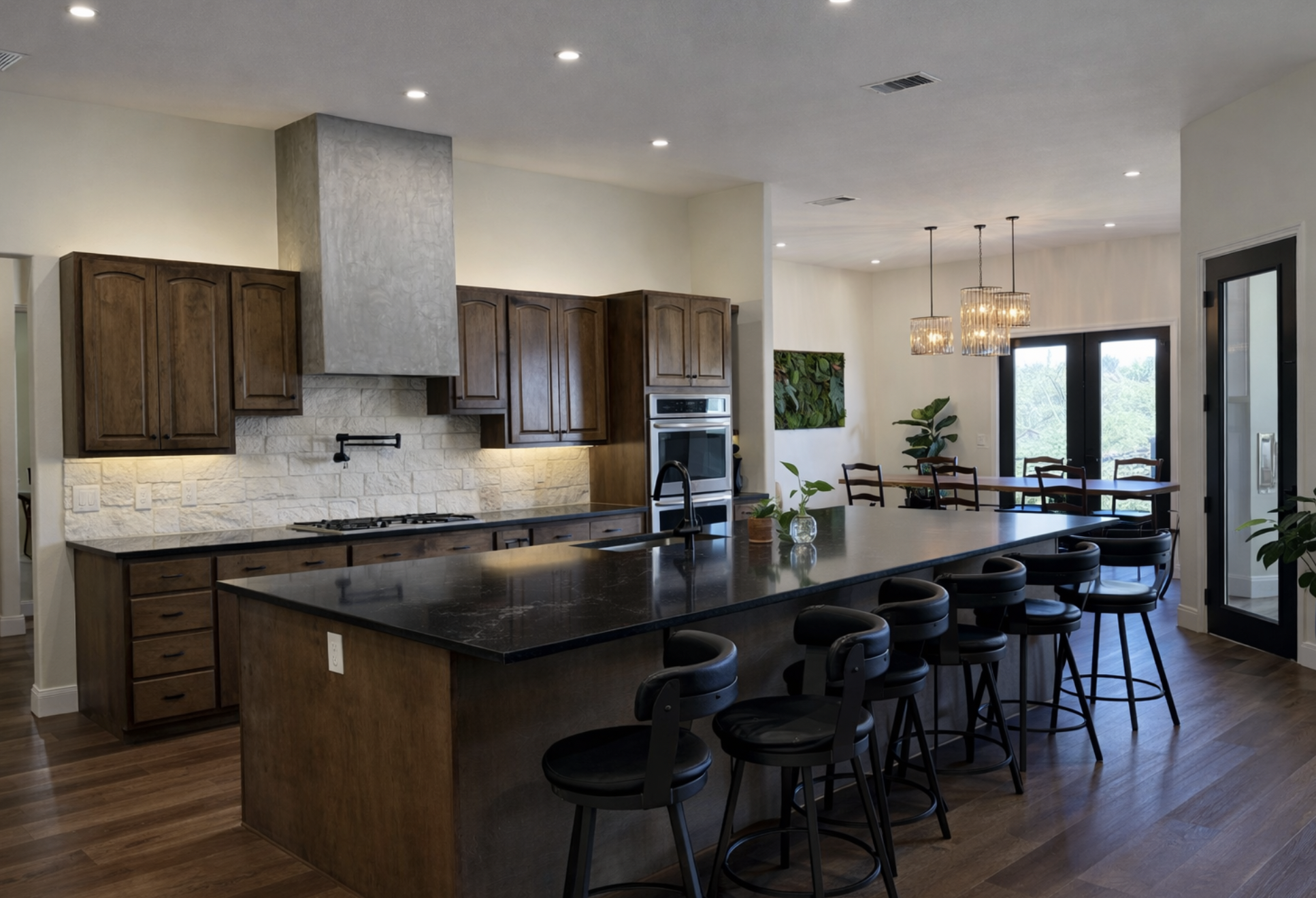 Custom built modern kitchen with wooden cabinets, black marble island counter, and dining area with hanging pendant lights and glass doors.