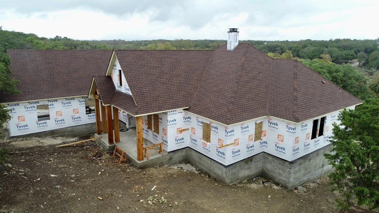 A house under construction with a brown shingled roof, wrapped in Tyvek HomeWrap, situated on a concrete foundation, surrounded by trees and construction materials.