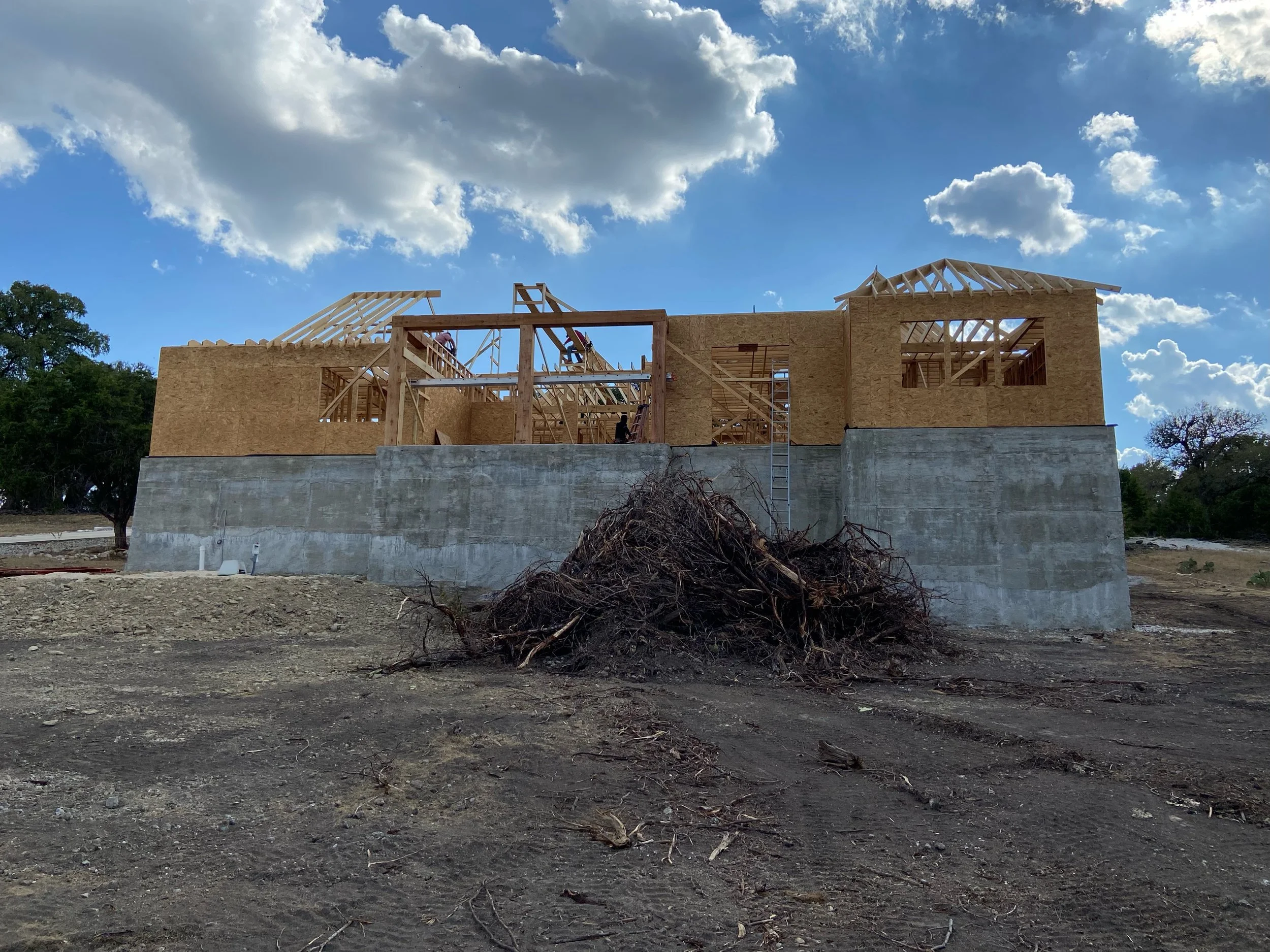 House under construction on elevated foundation with wooden framing and roofing trusses, dirt ground, some trees in background, partly cloudy sky.