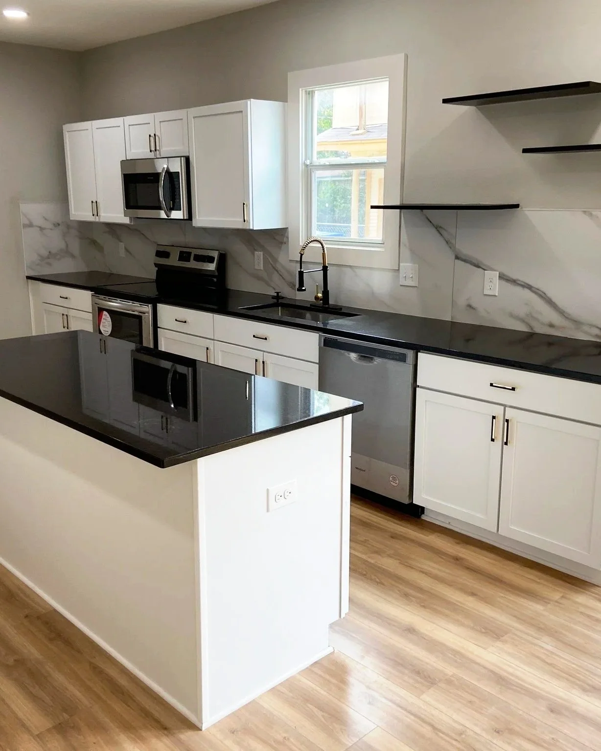 Modern kitchen with white cabinets, black countertops, a marble backsplash, stainless steel appliances, a window above the sink, and open shelving.