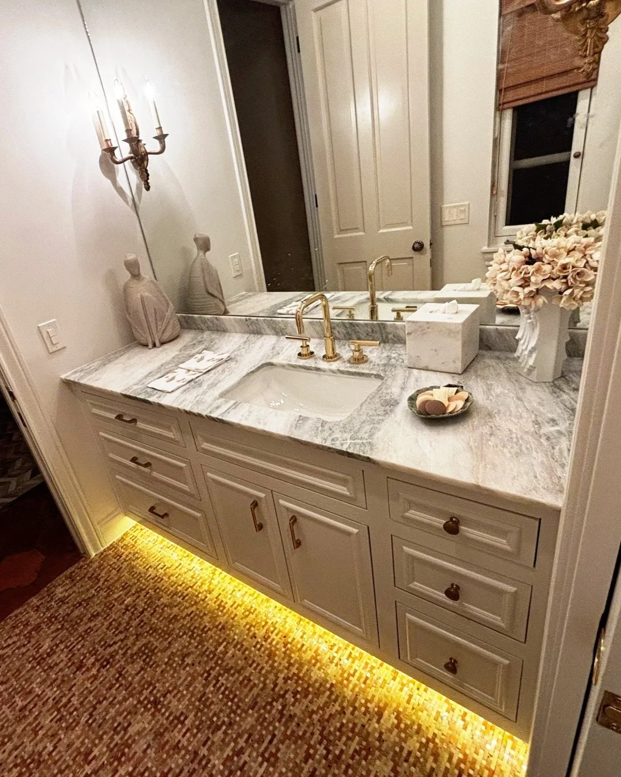 Bathroom vanity with a marble countertop, gold faucet, and a large mirror. Decor includes two beige ceramic figures, a vase with flowers, a tissue box, and a small dish with soap. The vanity has multiple drawers and cabinets with brass handles. There's warm yellow under-cabinet lighting and a glimpse of a patterned carpet.