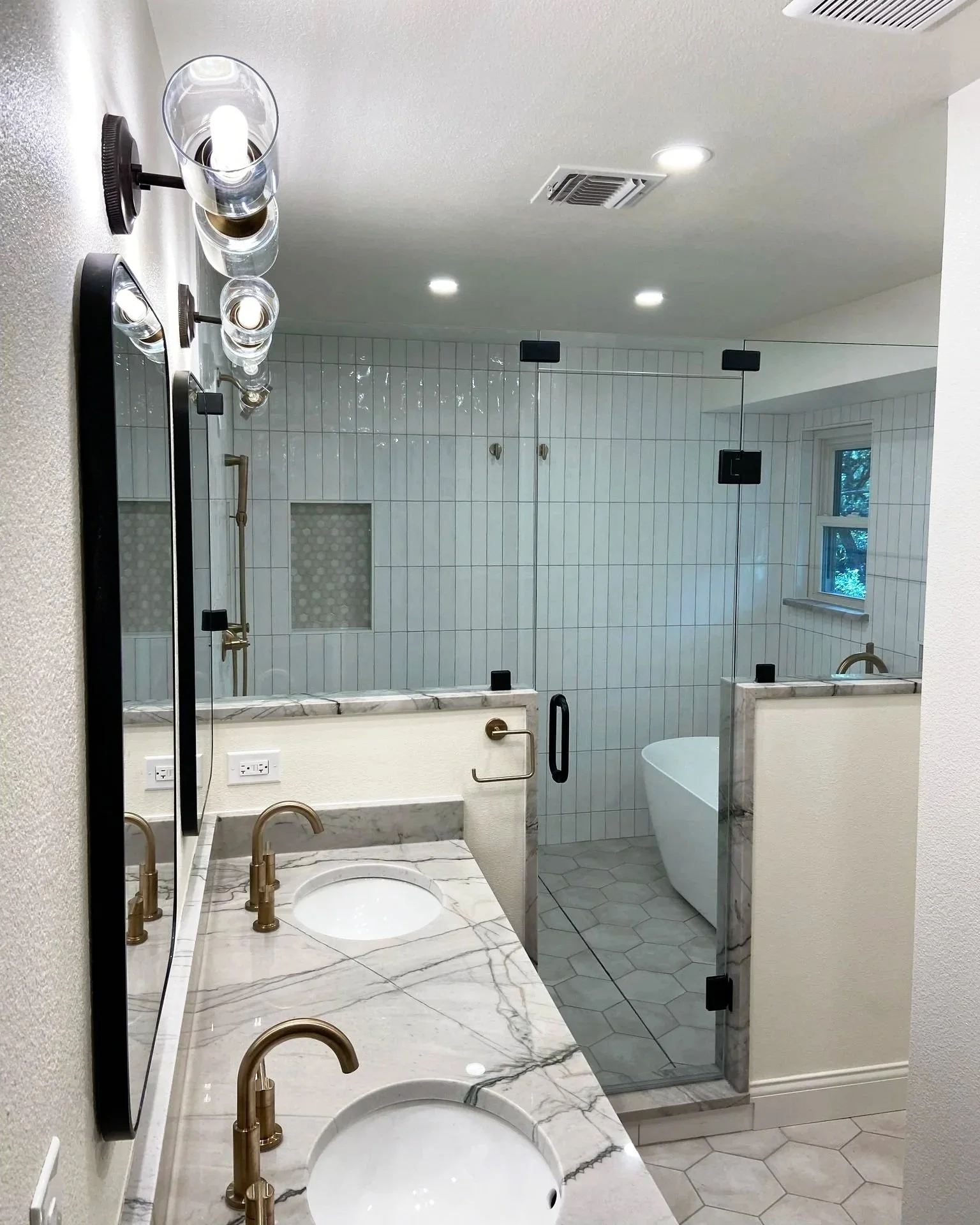 A modern bathroom with double marble sinks, a glass enclosed shower, and a freestanding bathtub. The bathroom has gold fixtures, a large mirror, and a window with a view of trees outside.