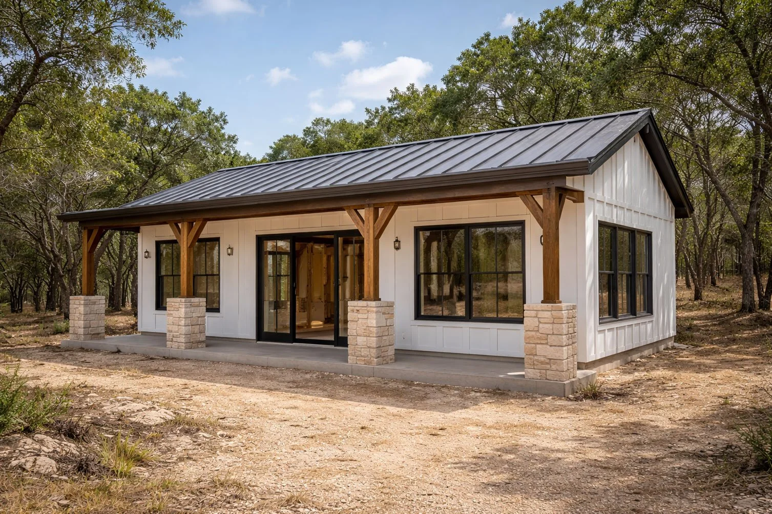 A modern single-story house with white exterior walls, black-framed windows, a metal roof, and a front porch with wooden beams supported by stone columns, set in a wooded area under a partly cloudy sky.