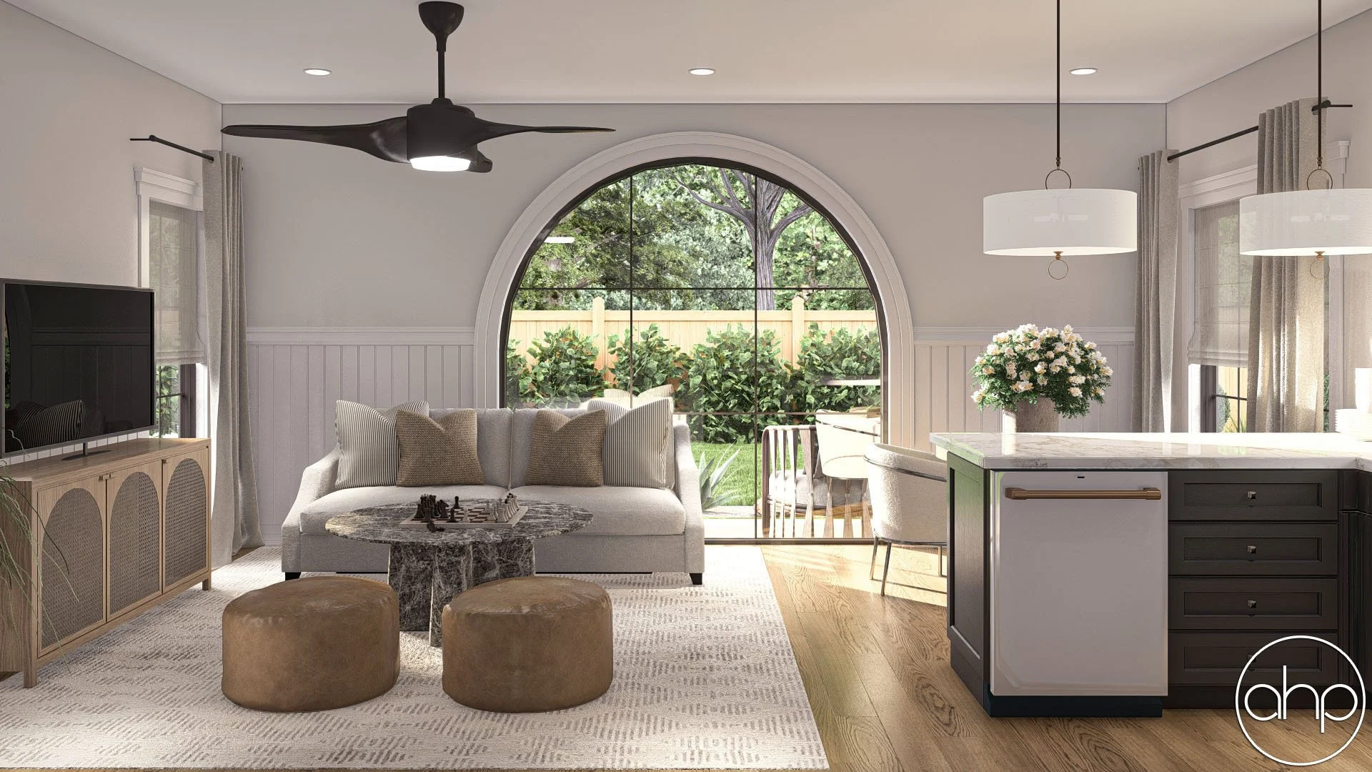 Modern living room with large arched window showing a backyard garden, featuring a gray sofa, two round ottomans, a marble coffee table with a chessboard, a TV on a wooden stand, white and beige pillows, greenery and outdoor furniture visible through the window, and a kitchen counter with a flower arrangement.