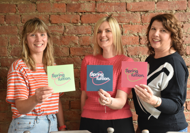 Three women standing in front of a brick wall, each holding a sign that says 'Spring Tuition' in different colors.