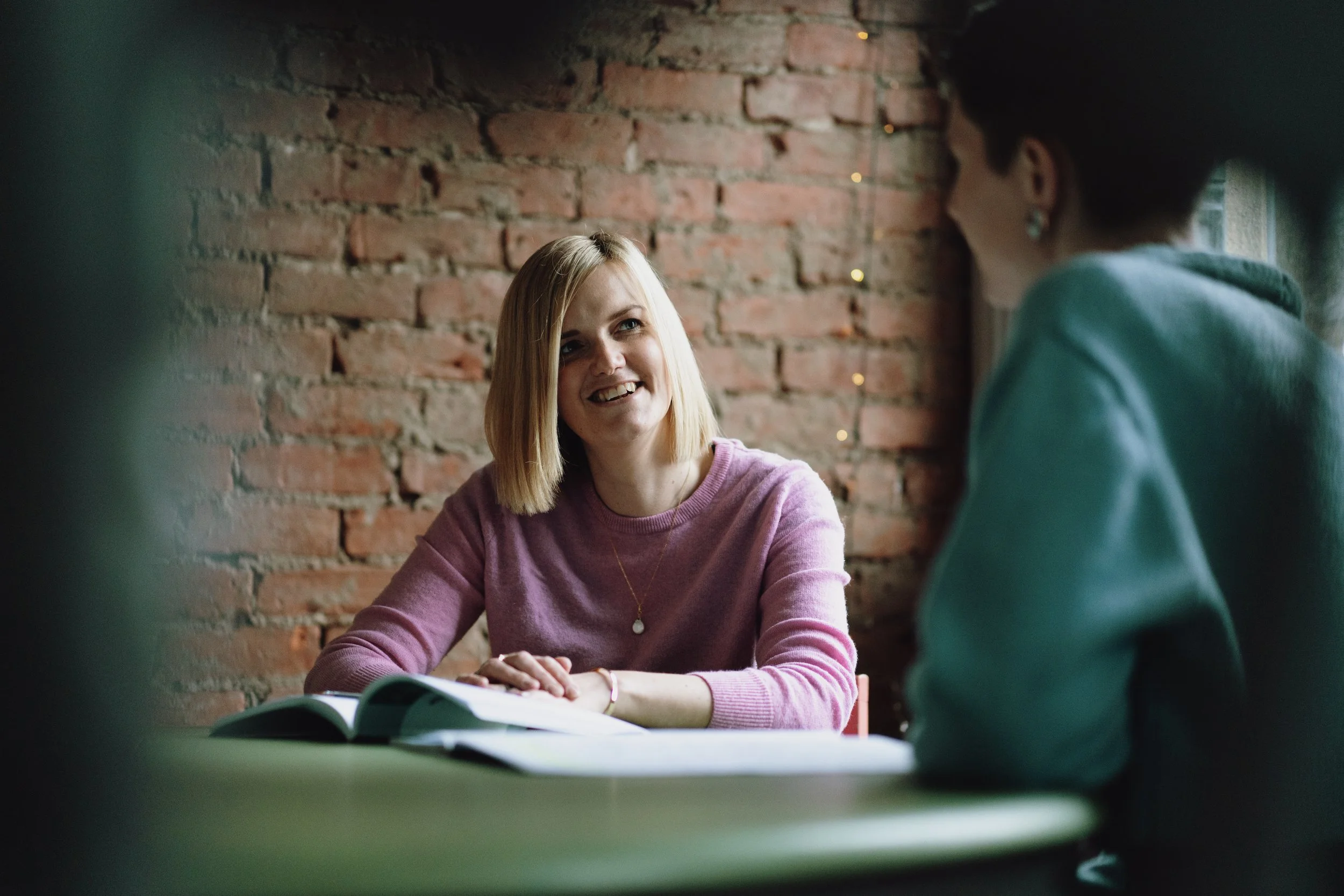A woman with blonde hair and a pink sweater smiling while talking to a person with short hair, wearing earrings, sitting in front of her against a brick wall.