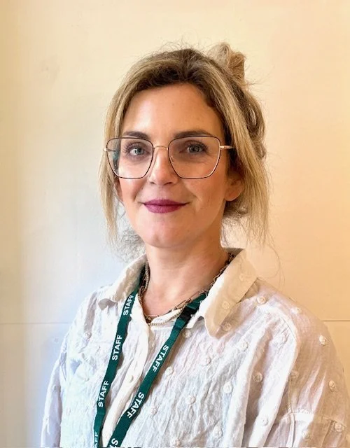 A woman with glasses and wavy light brown hair pulled back, wearing a white shirt and a green lanyard that says 'STAFF.'