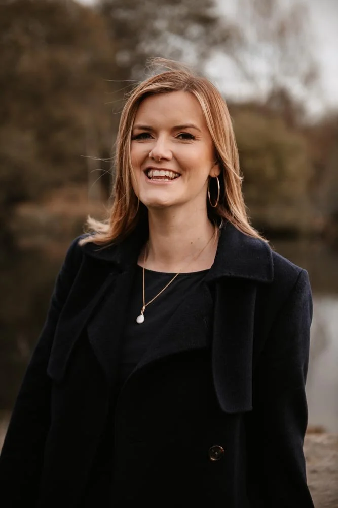 A smiling woman with shoulder-length blonde hair, wearing hoop earrings, a necklace, and a black coat outdoors with blurred trees and water in the background.