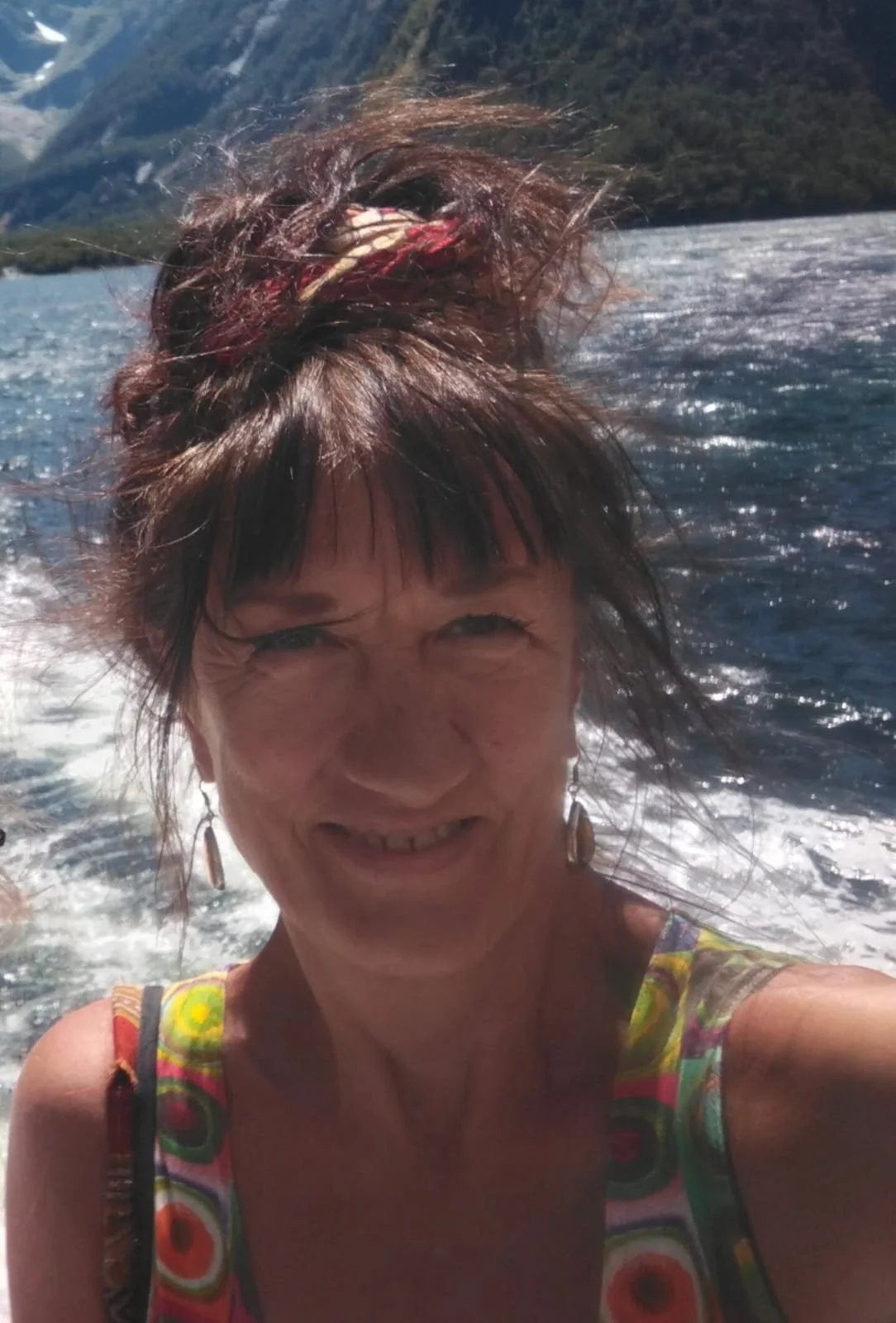 A woman with short dark hair, wearing earrings and a colorful patterned dress, smiling at the camera with water and mountains in the background.