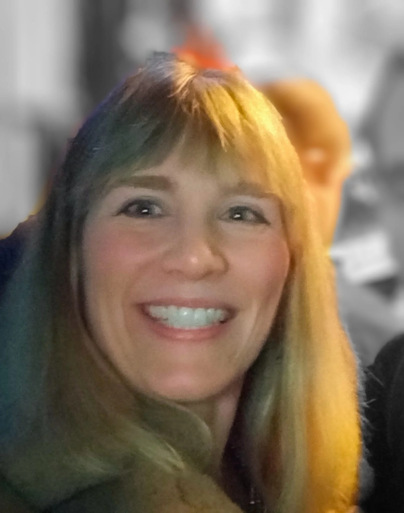 A close-up of a woman smiling, with blond and dark hair, wearing a yellow jacket.