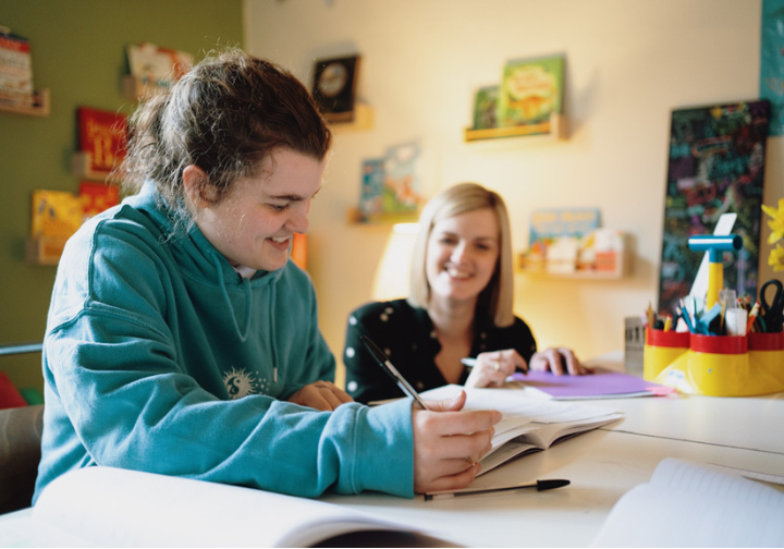 Two young women sitting at a table engaged in a homework or study session with books, notebooks, and school supplies, in a colorful room with shelves of books and decorations.