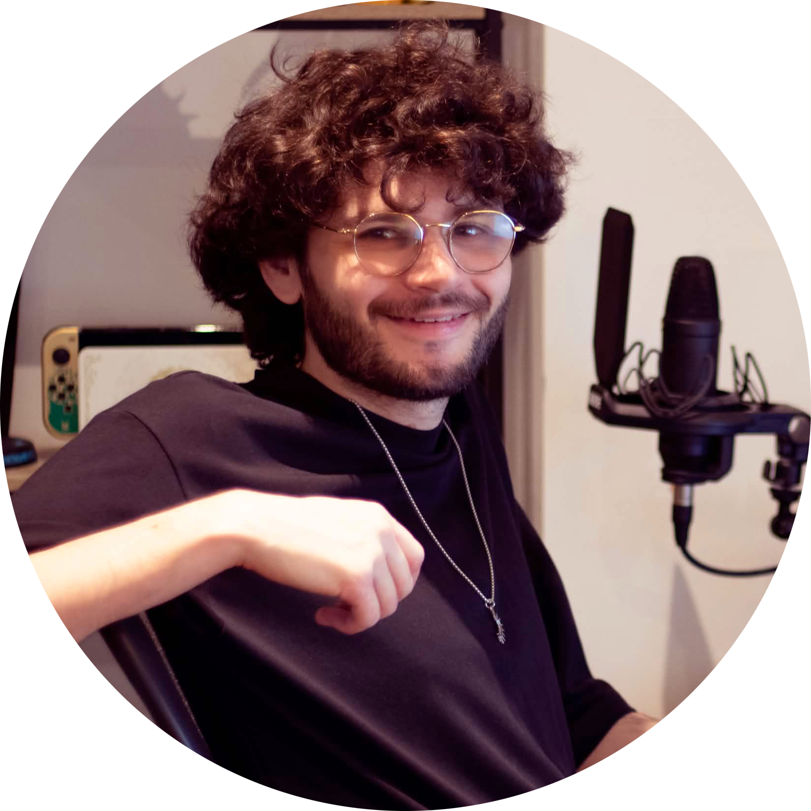 A young man with curly hair, glasses, and a beard smiling in with a microphone in front of him.