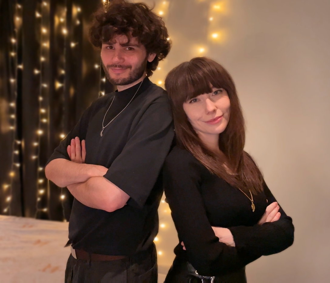 A young man and woman standing back-to-back with crossed arms, smiling at the camera, in front of a wall with warm string lights.
