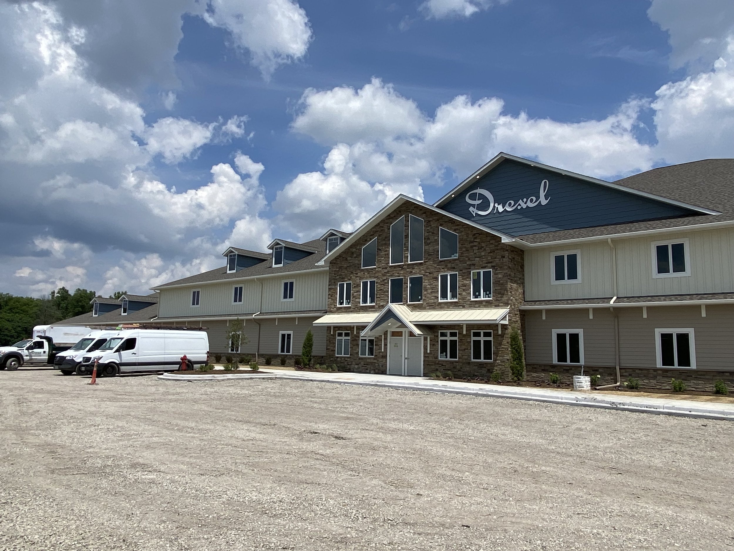 New multi-story apartment building under a partly cloudy sky with a gravel parking lot and vehicles in front.