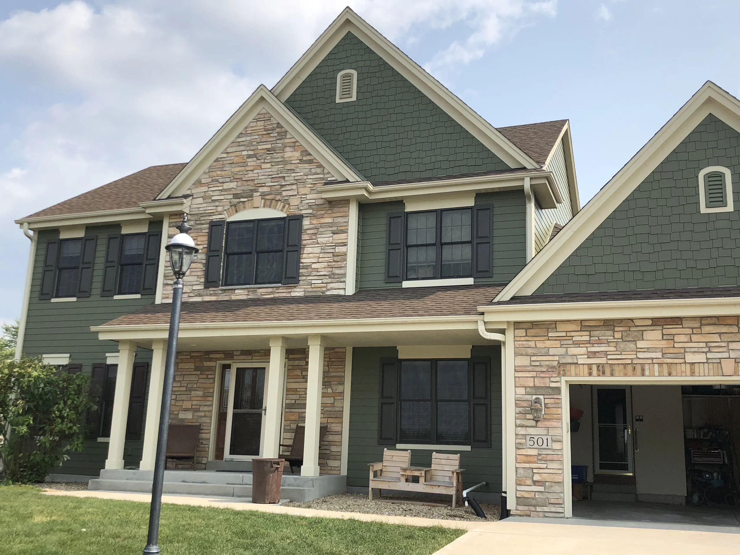 A modern two-story house with green siding, stone accents, black shutters, and a driveway leading to an open garage.