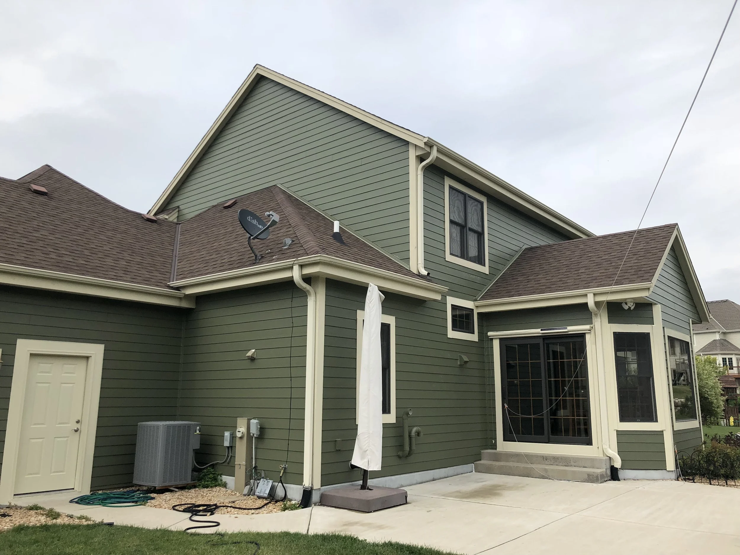 Back of a two-story house with green siding, brown shingle roof, satellite dish, air conditioning unit, and patio umbrella, with concrete patio and steps.