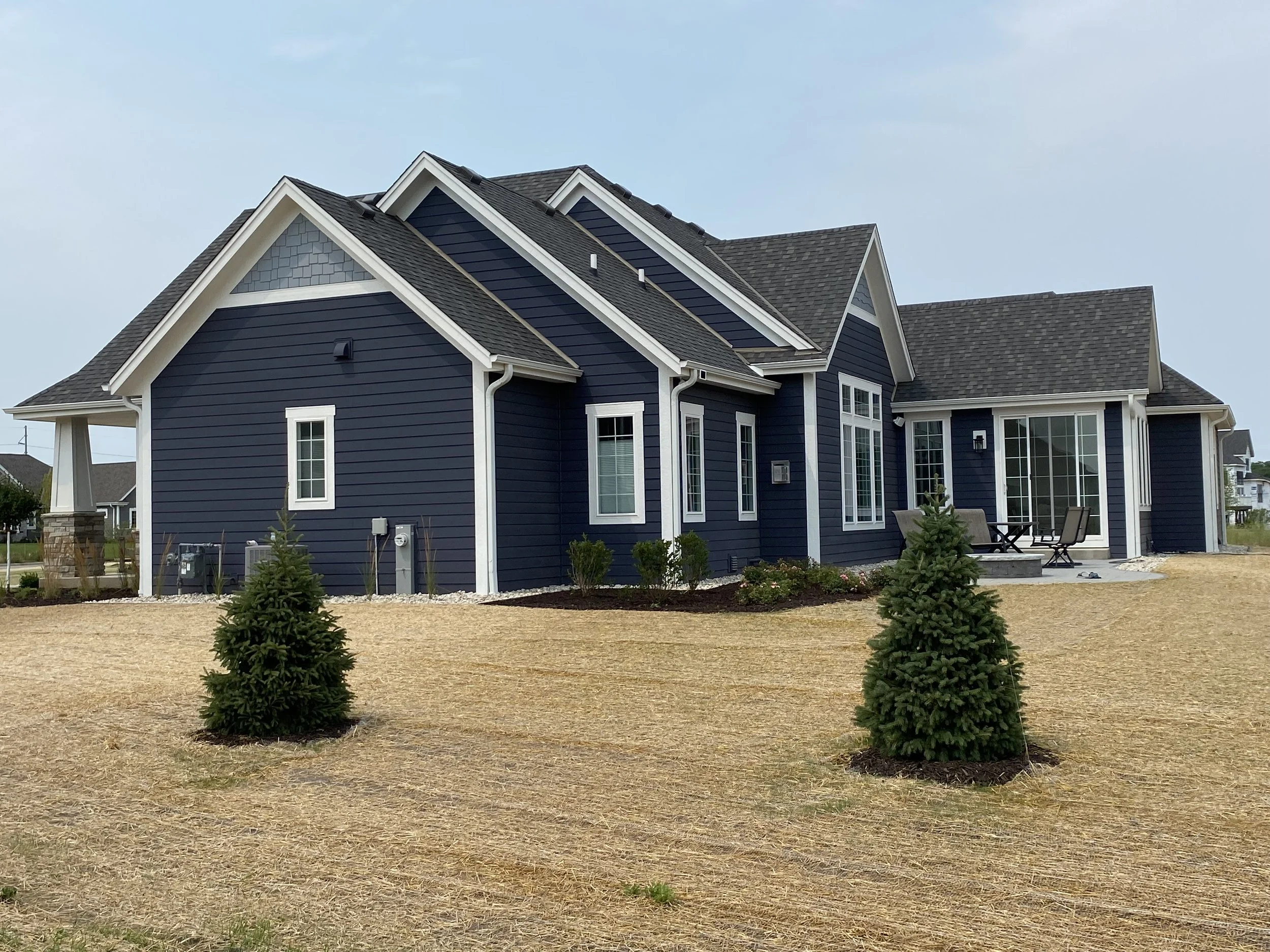 A blue house with white trim and multiple gabled roofs, surrounded by small evergreen trees and a landscaped yard with gravel and mulch.