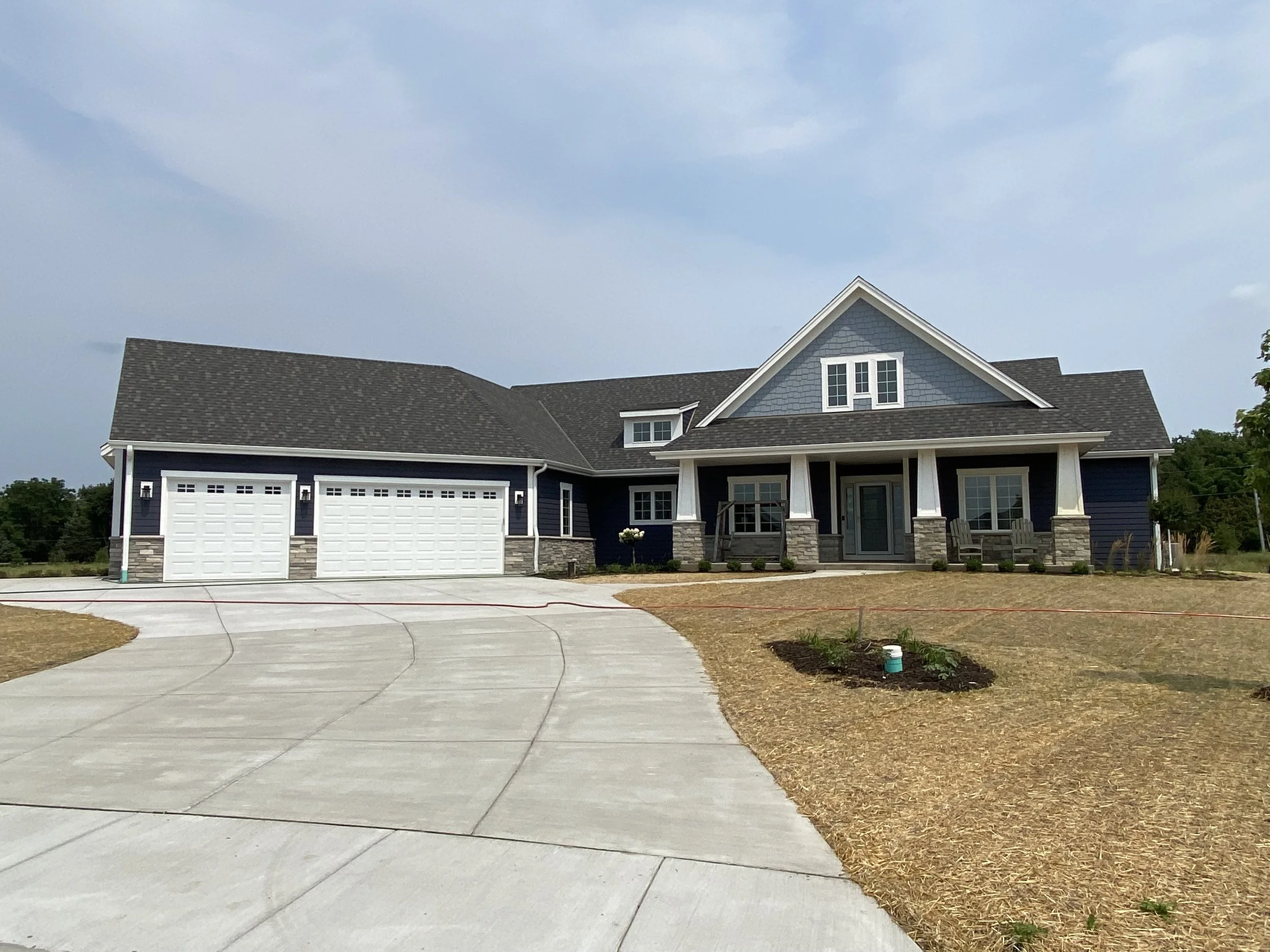 A large, modern house with dark blue siding, white trim, and a stone foundation. It has a three-car garage, a covered front porch with pillars, and a gabled roof. The yard is partially landscaped with freshly planted shrubs and flowers, and a wide co