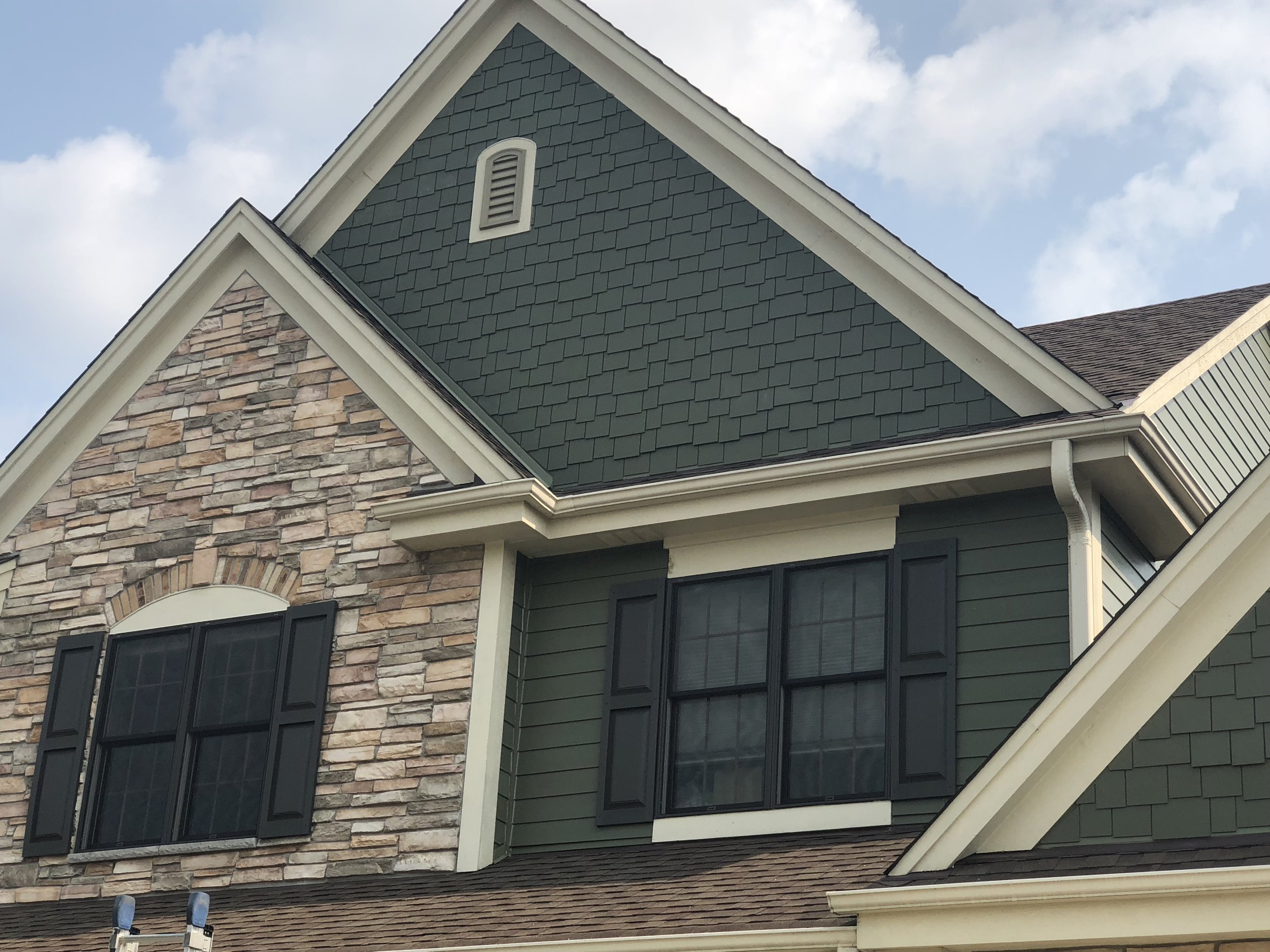 Close-up view of the upper part of a house with a mix of stone and siding exterior, two black window shutters, and a gable roof with clouds in the sky.