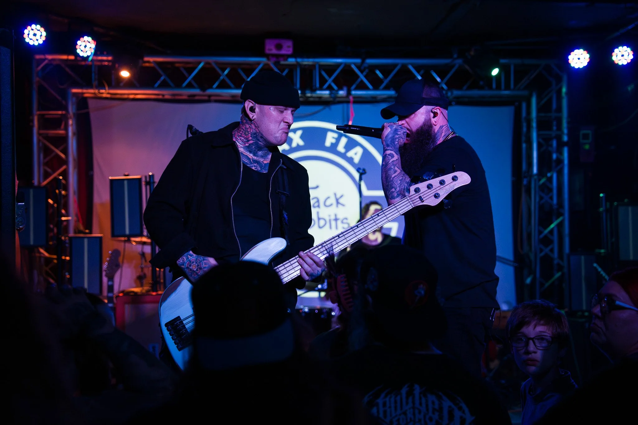 Two tattooed male musicians performing on stage; one playing bass guitar, the other singing into a microphone. Stage lighting is colorful with a logo in the background that reads 'Black FLA'. Audience members are visible in the foreground.