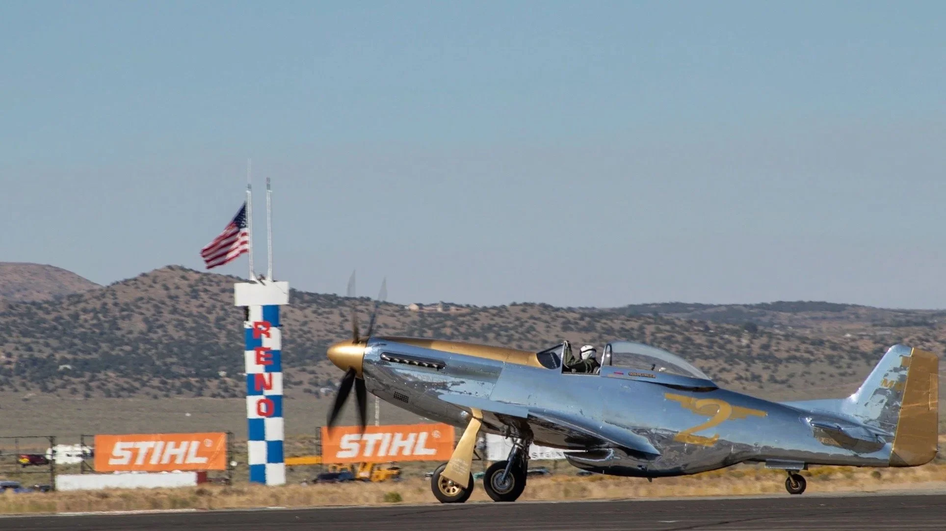 A silver and gold vintage aircraft taxiing on the runway with an American flag waving, and Stihl sponsor banners in the background.
