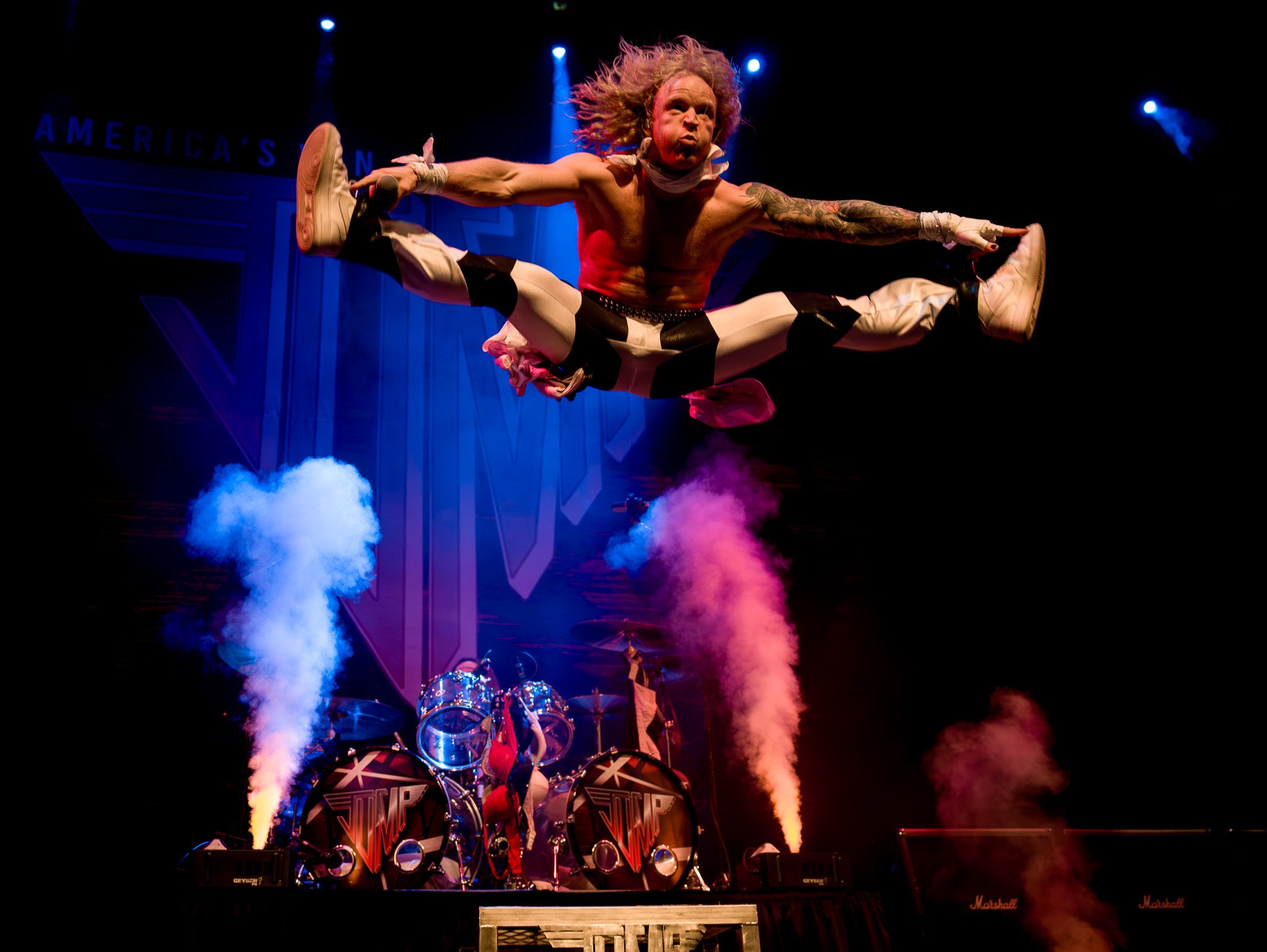 Performing musician with long curly hair jumping mid-air on stage, wearing black and white pants and sneakers, with a backdrop reading 'America's'. Stage smoke and drums are visible below.
