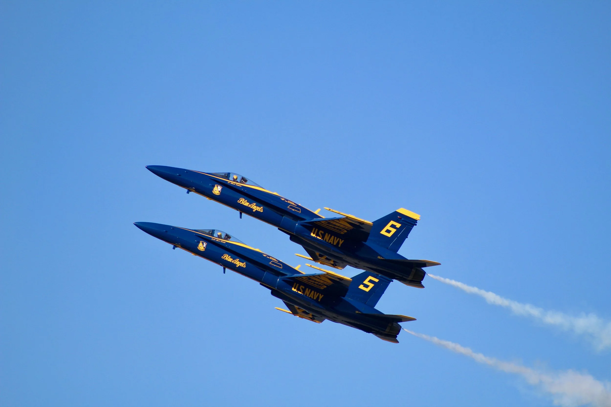 Two U.S. Navy Blue Angels jets flying in formation against a clear blue sky, leaving white smoke trails behind.