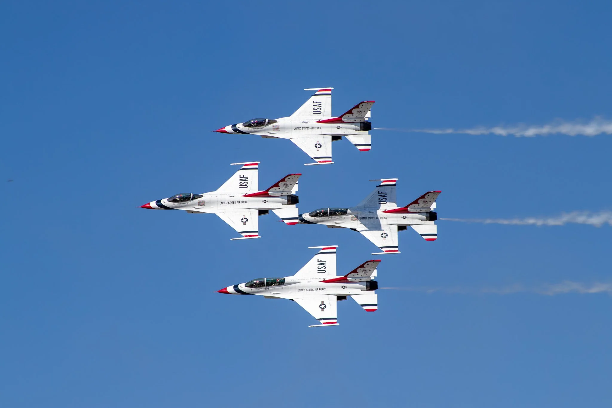 Four United States Air Force fighter jets flying in a coordinated arrow formation against a blue sky, with vapor trails behind them.