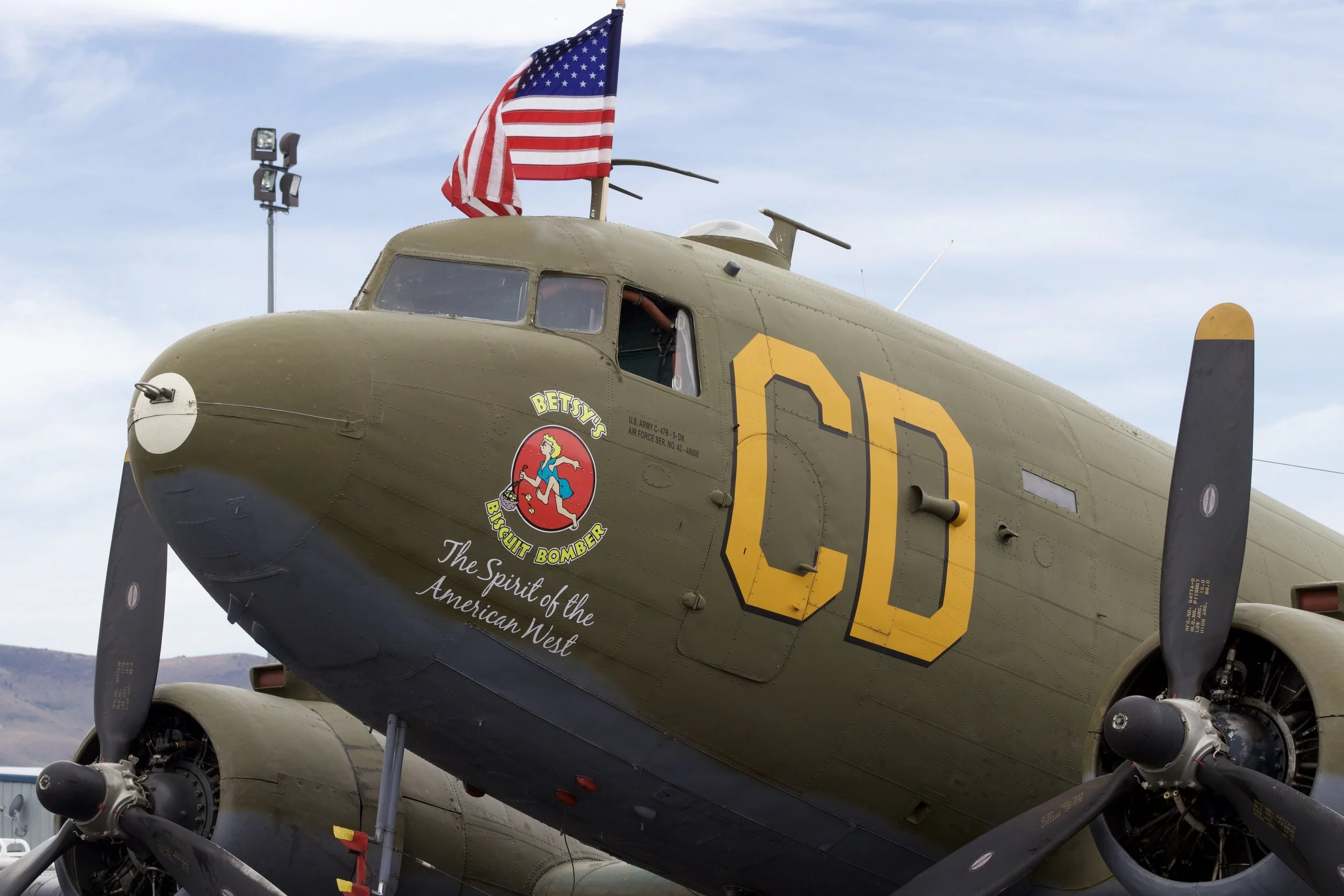 A vintage military aircraft with an American flag flying behind it. The plane is painted green and has large yellow letters 'CD' on its side. There is a cartoon emblem of a girl with blonde hair running, labeled "Betsy the Biscuit Bomber," and the te