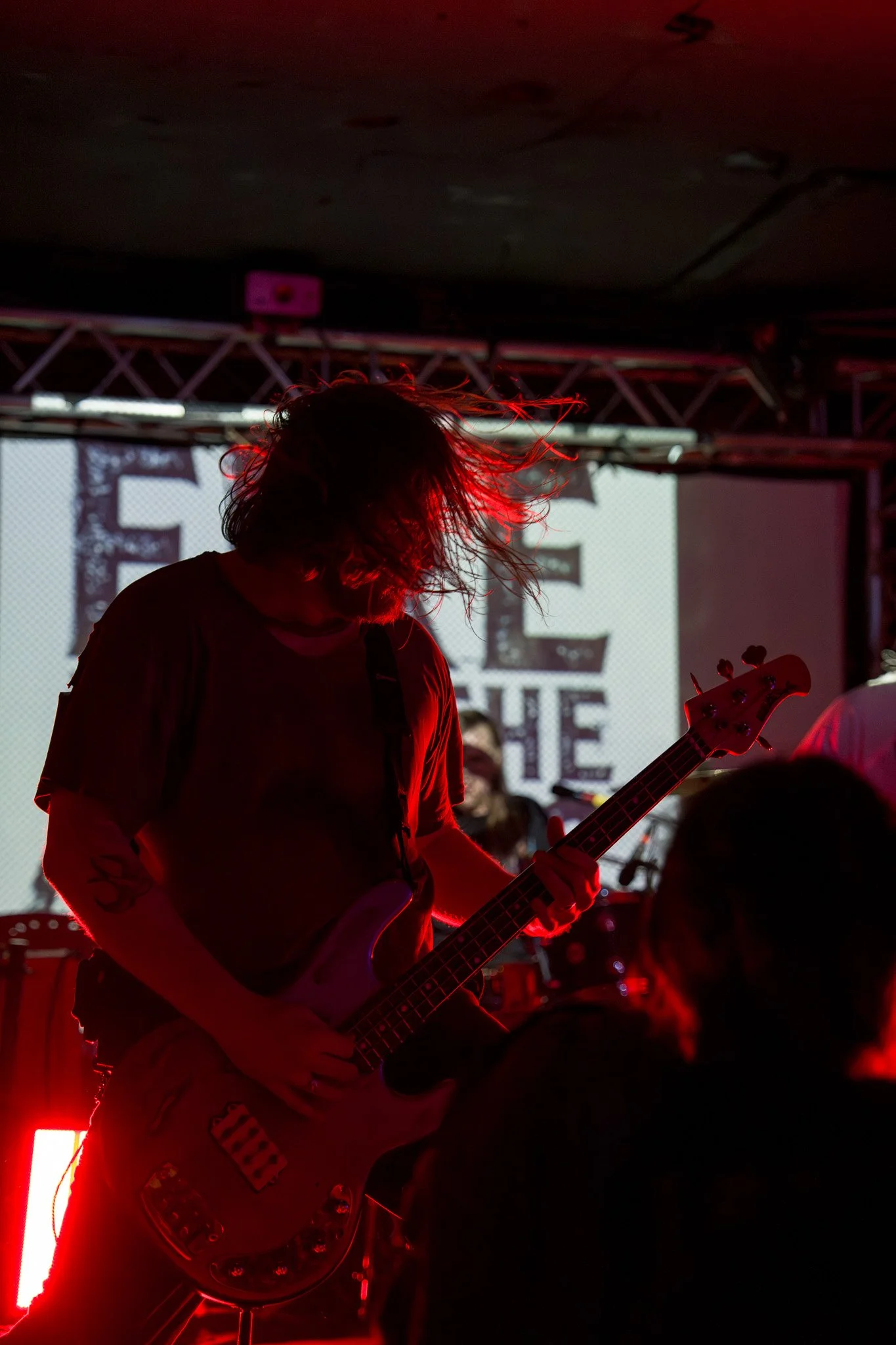 A musician playing an electric guitar on stage, backlit with red lighting, with their hair swinging as they move.