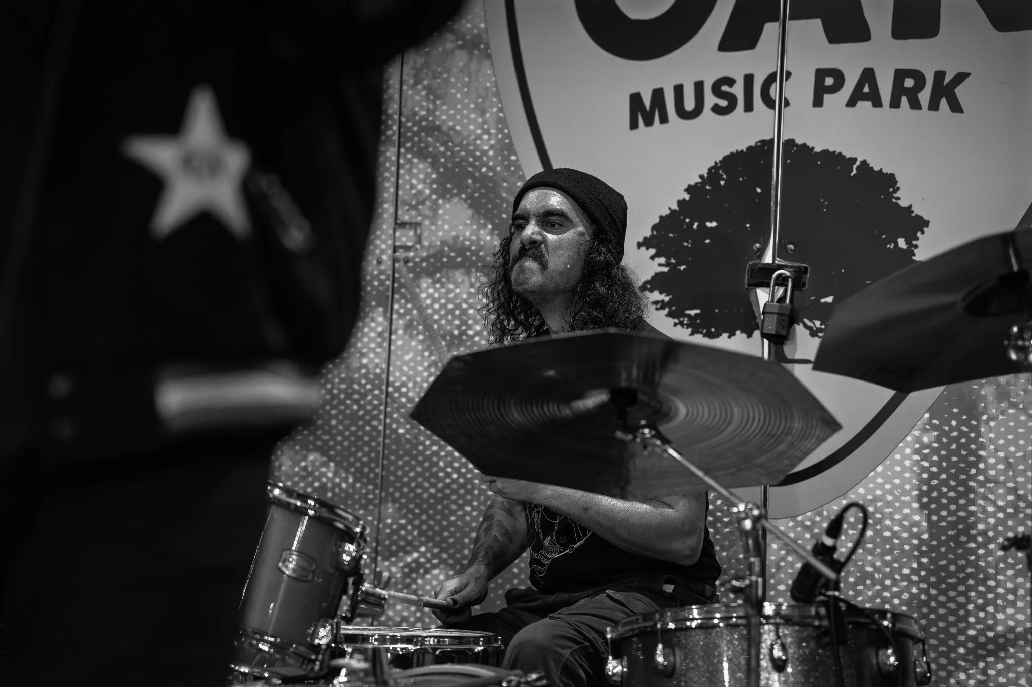 A musician with long, curly hair, a beard, and a beanie playing the drums at a music park, with a sign reading 'Music Park' and a tree in the background.