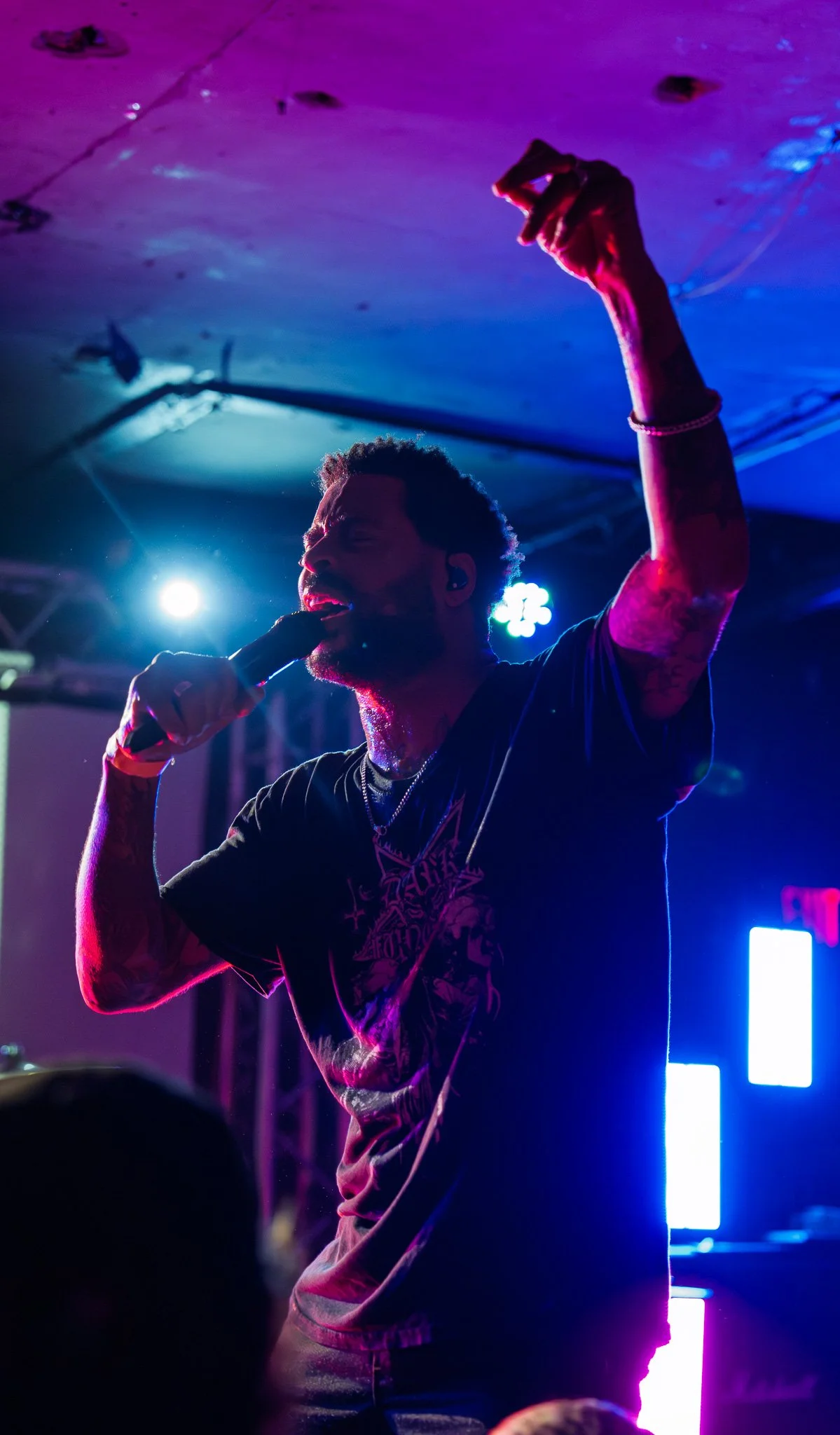 A singer with tattoos on his arms singing into a microphone during a concert, illuminated by colorful stage lights.