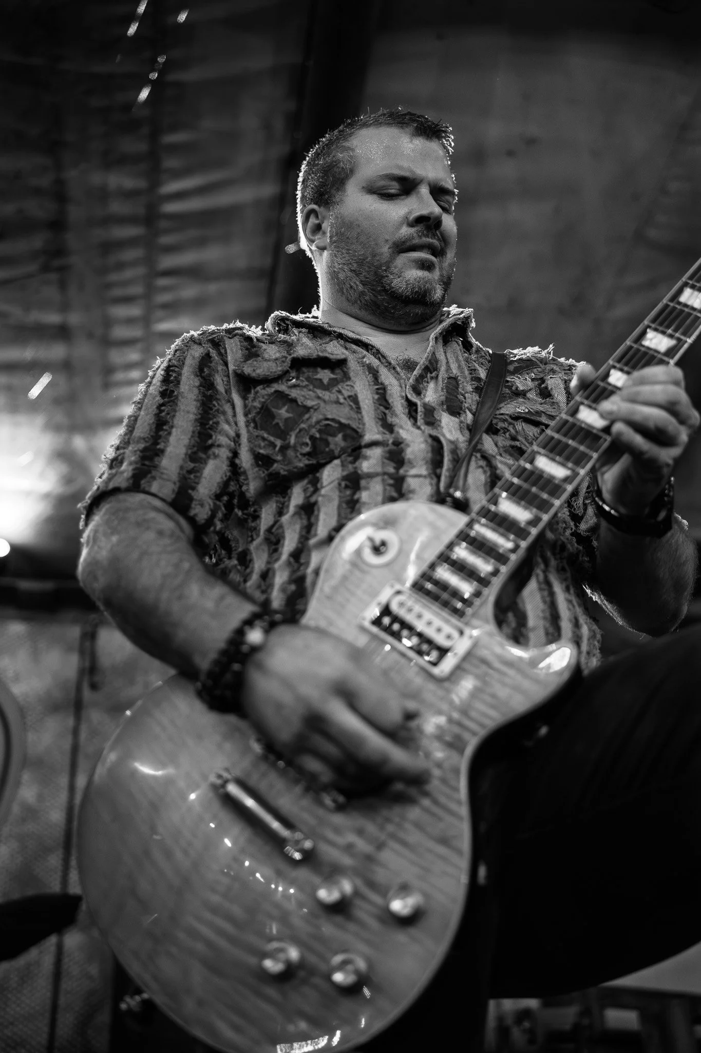 A man playing an electric guitar indoors, wearing a distressed American flag shirt, with a focused expression.