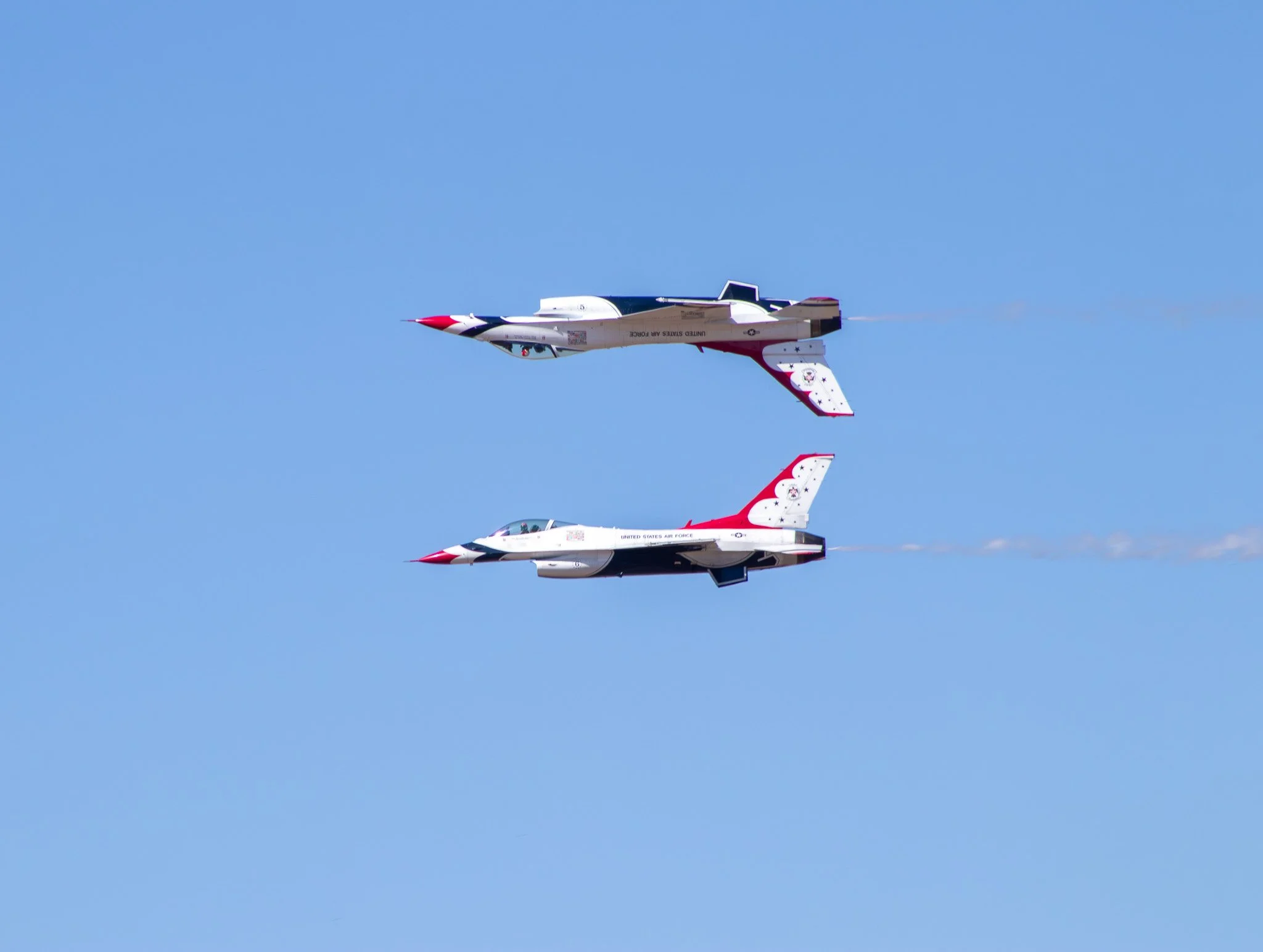 Two fighter jets performing a mirror image flying maneuver against a blue sky, with smoke trails visible behind them.