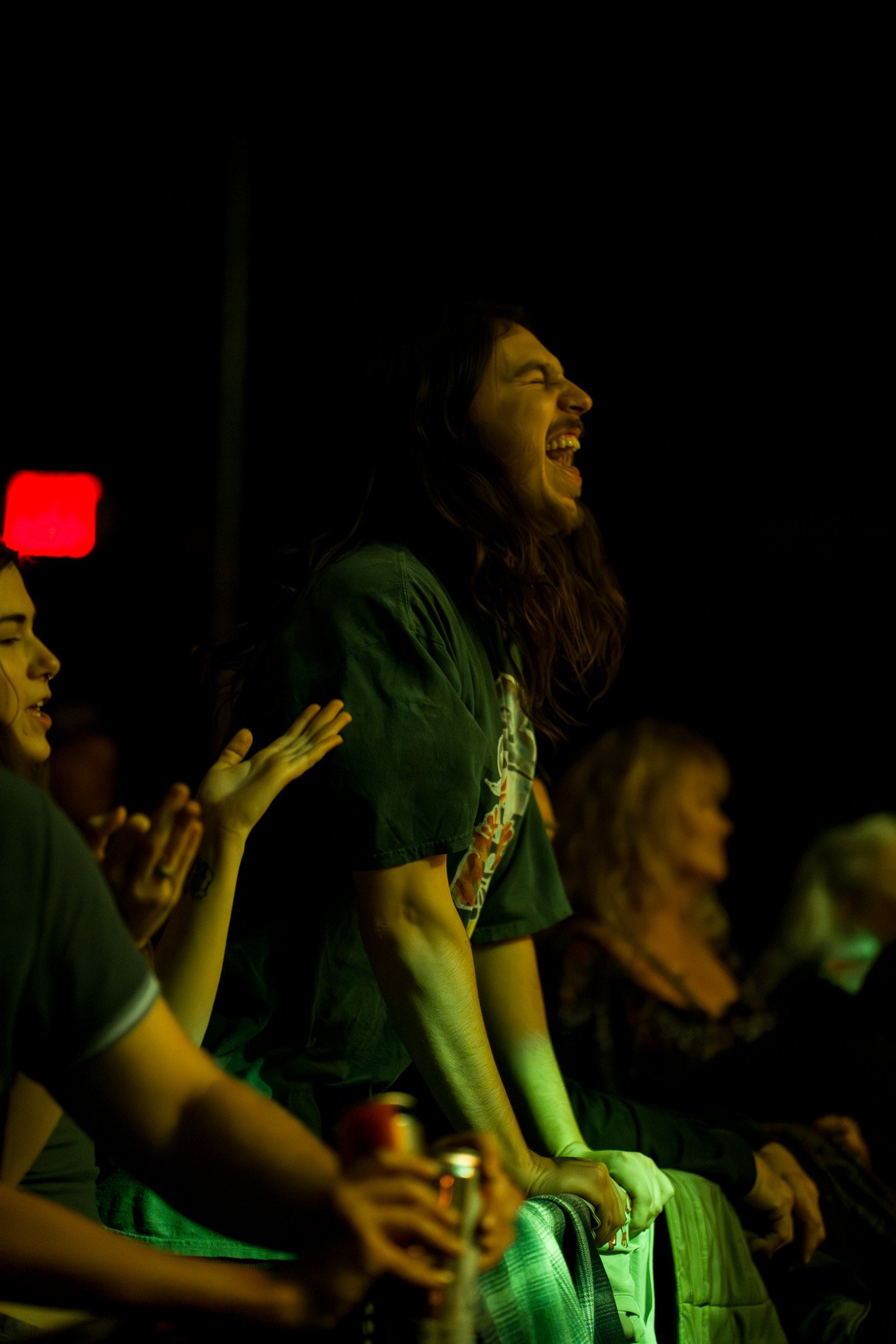 A group of people at a concert or event, with a woman in the center laughing with her eyes closed, wearing a green shirt, illuminated by green lighting.