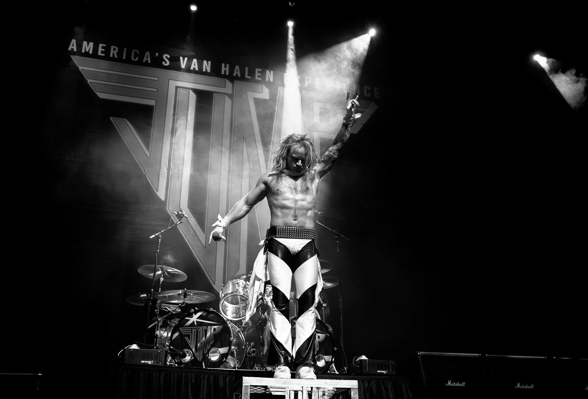 A black and white photo of singer on stage during a concert, with a large 'Vamp' logo and 'America's Van Halen Experience' banner in the background. The singer has long blonde hair, is shirtless, wearing patterned pants, and holding a microphone whil