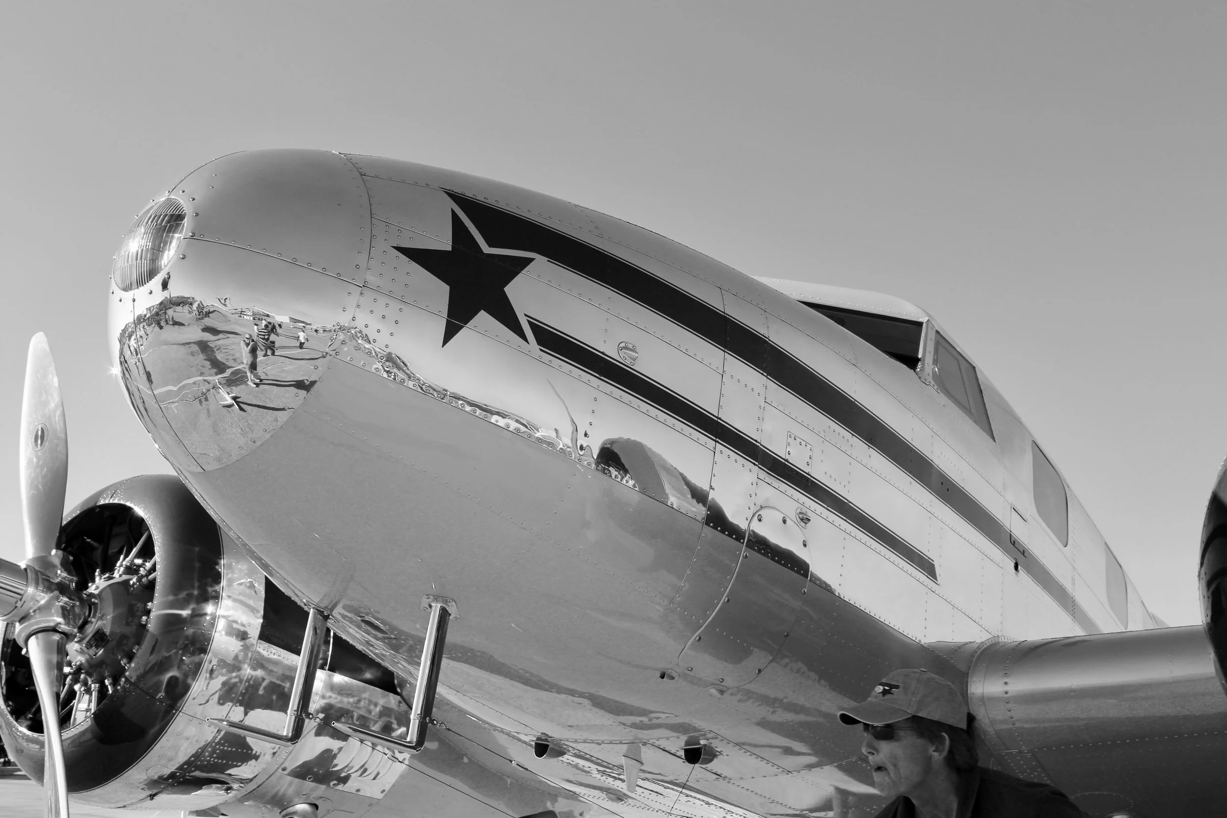 A vintage aircraft with a reflective chrome finish and black star decal, parked at an airshow with people visible through its reflection.