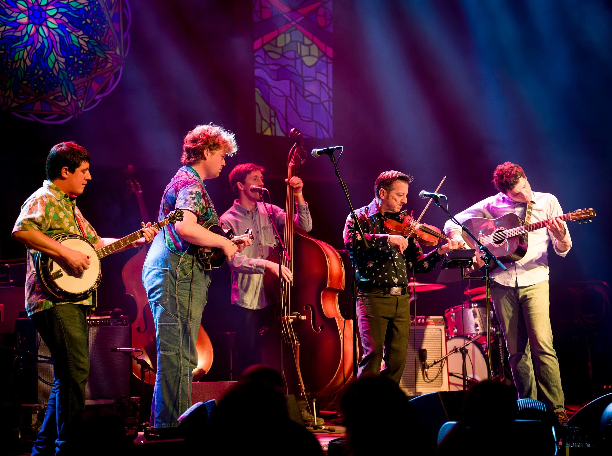 Group of six musicians playing instruments on stage during a performance, with colorful stained glass windows in the background and stage lighting.