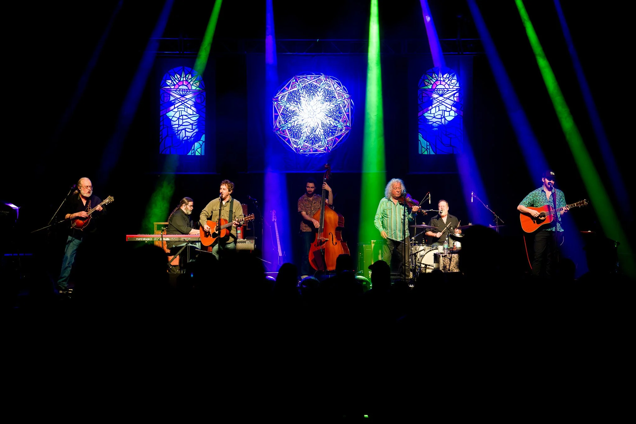 A band of seven musicians performing on stage with colorful lights and stained glass window background.