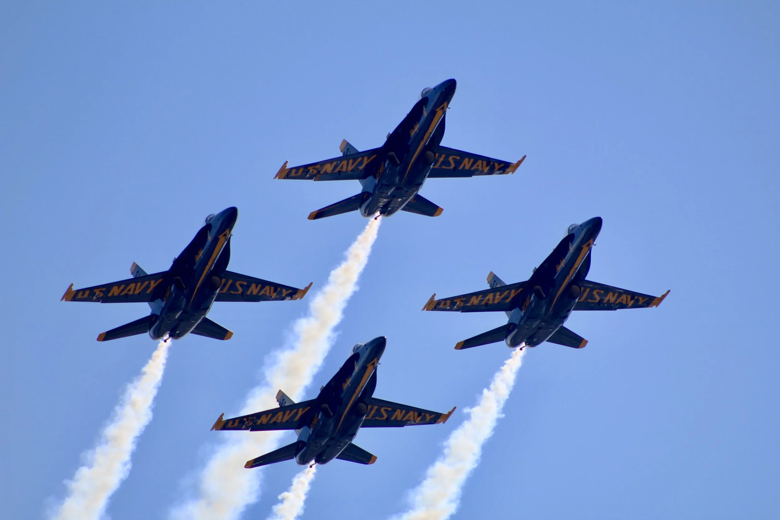 Four US Navy fighter jets flying in close formation, leaving white smoke trails against a clear blue sky.