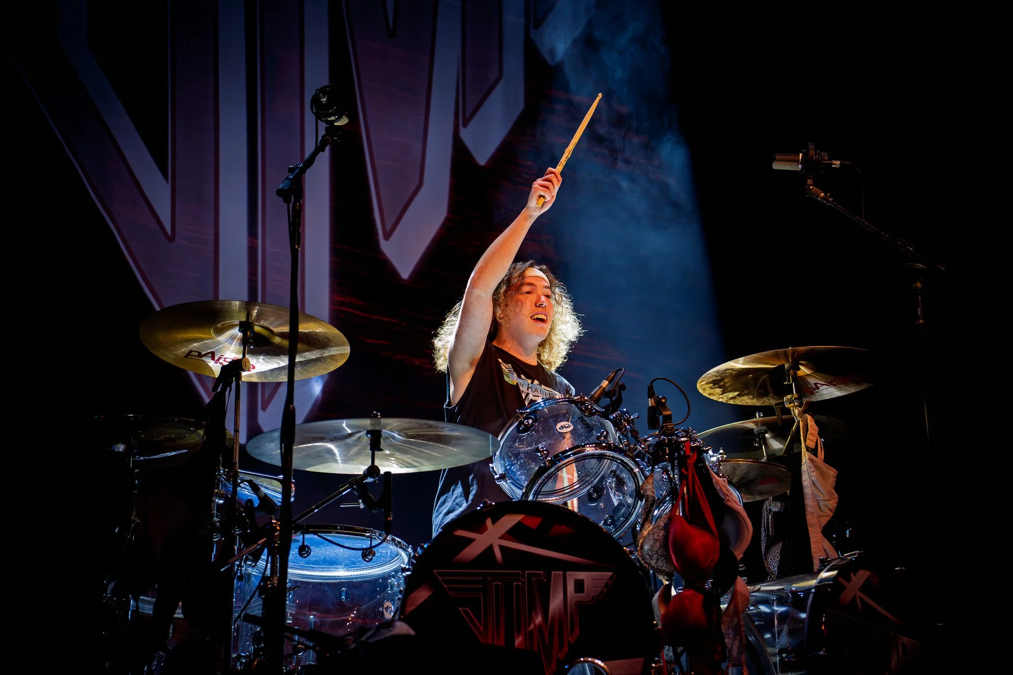 A drummer performing on stage with a drum set, raising drumsticks, illuminated by stage lights, and a large backdrop behind her.